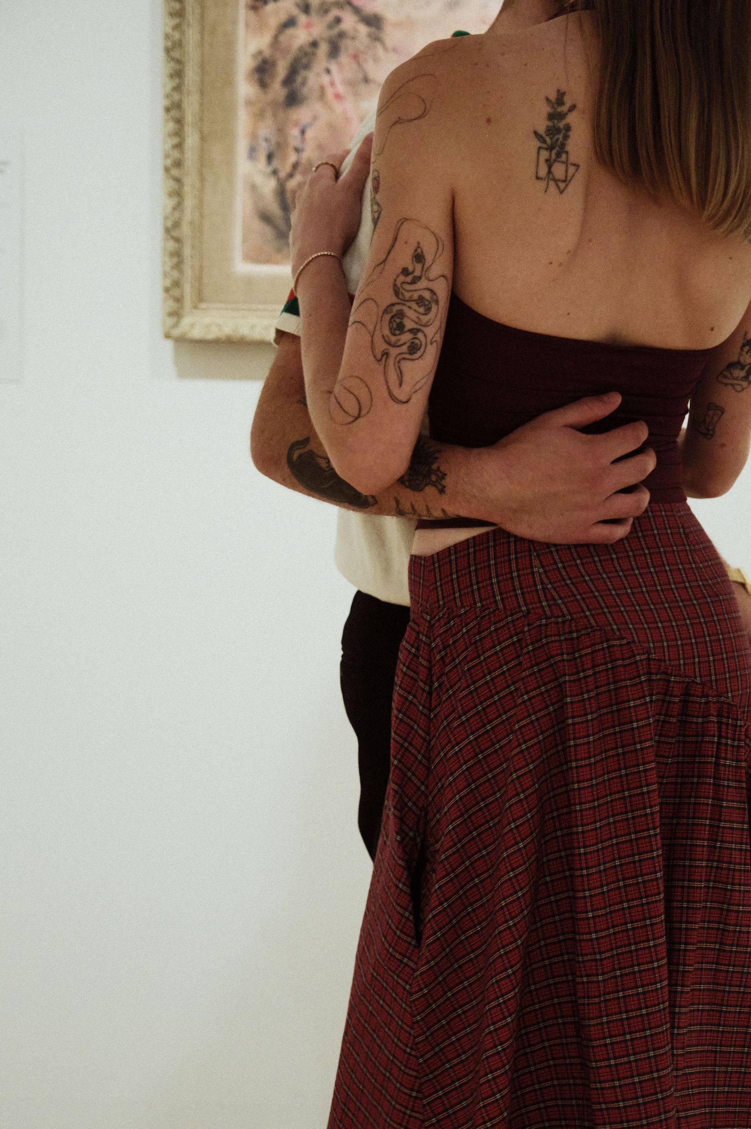 Couple embracing showing tattoos, woman wearing dark tube top and red plaid skirt in art gallery.