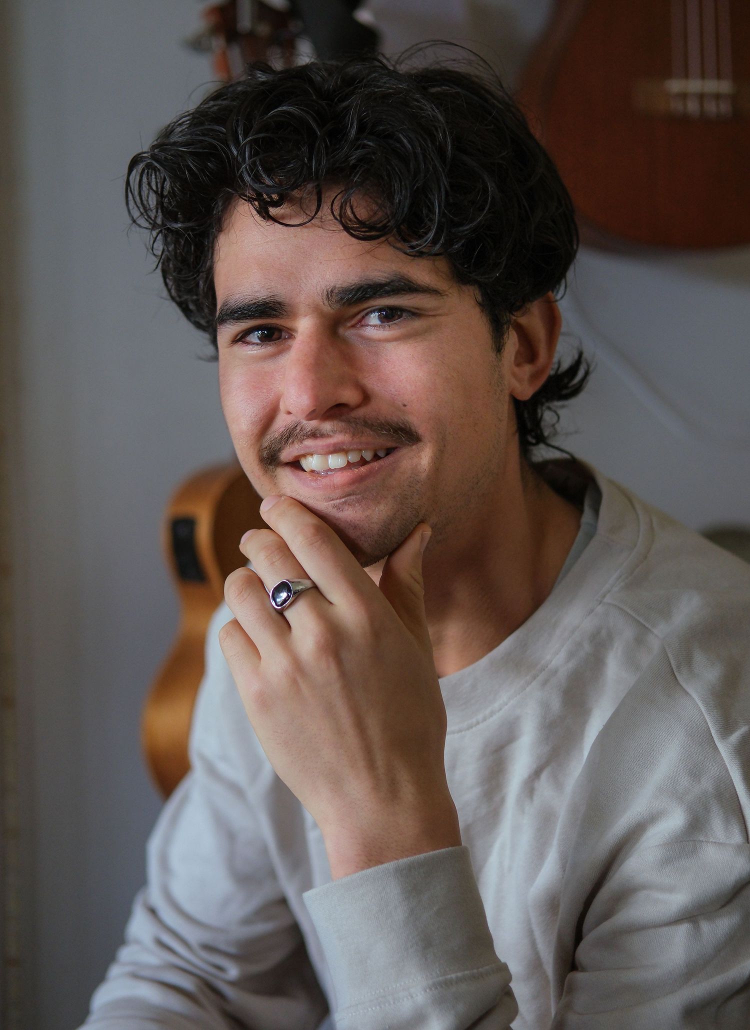 Smiling young man with curly hair and mustache posing with guitars on wall behind documentary style photography Visuals by Dani.