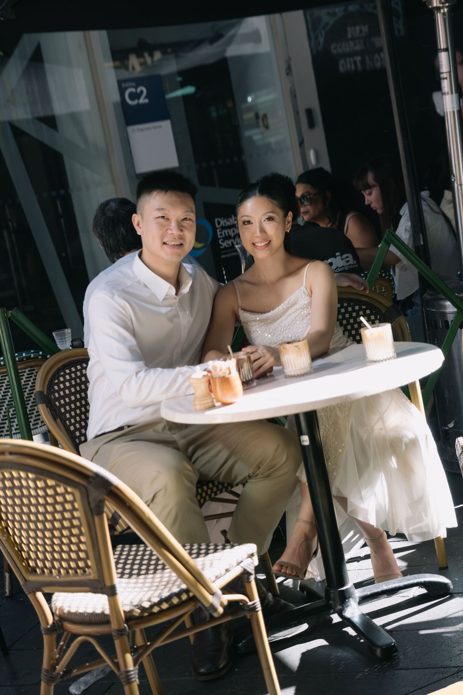 A couple smiles at an outdoor cafe table with coffee drinks, dressed in elegant white and beige attire.