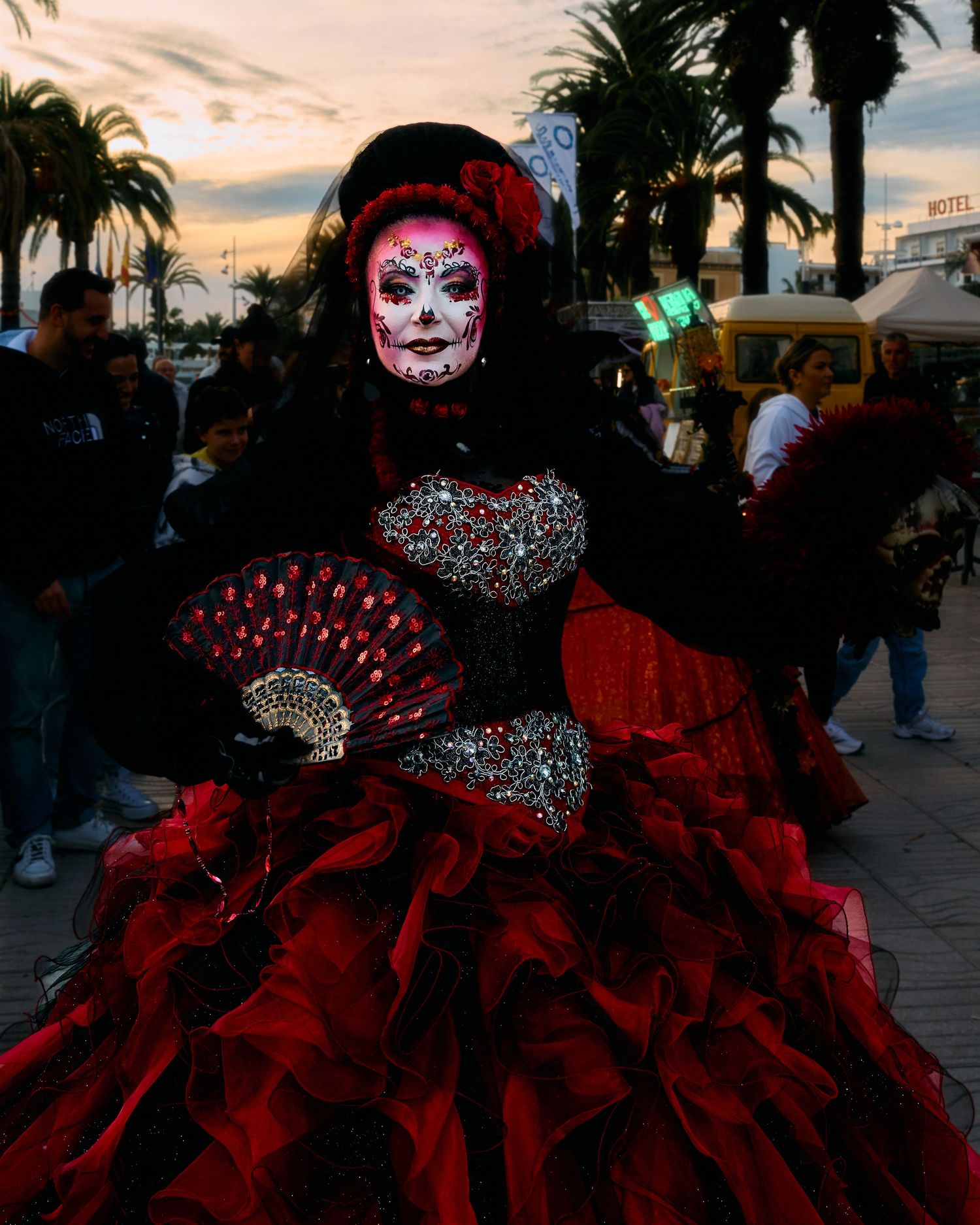 A person in Day of the Dead makeup and elaborate red dress holding a decorative fan at dusk.