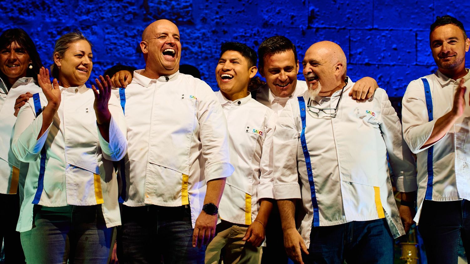 Group of professional chefs posing together against a bright blue backdrop.