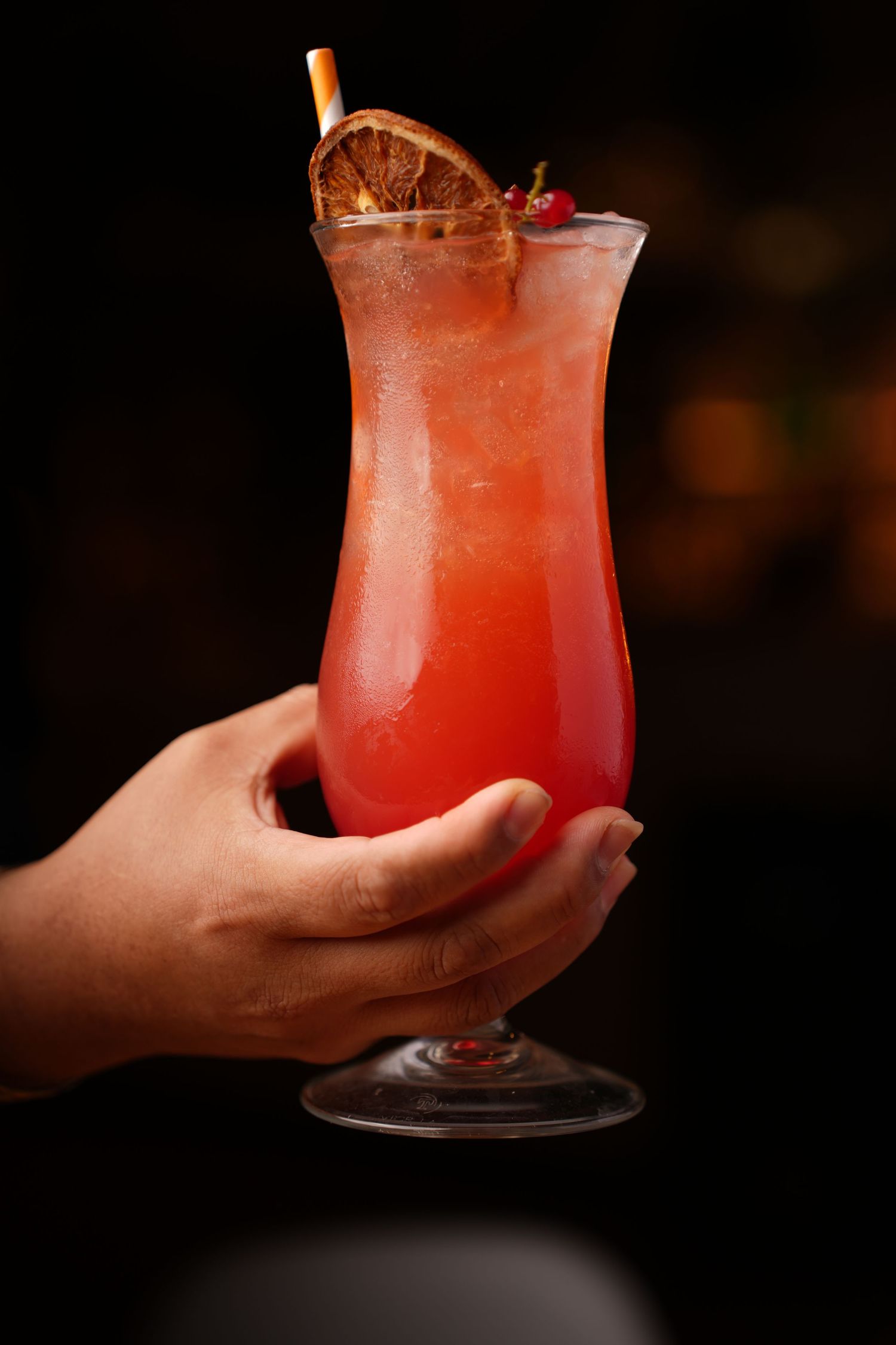 Tropical cocktail with orange garnish in tall glass held against dark background.
