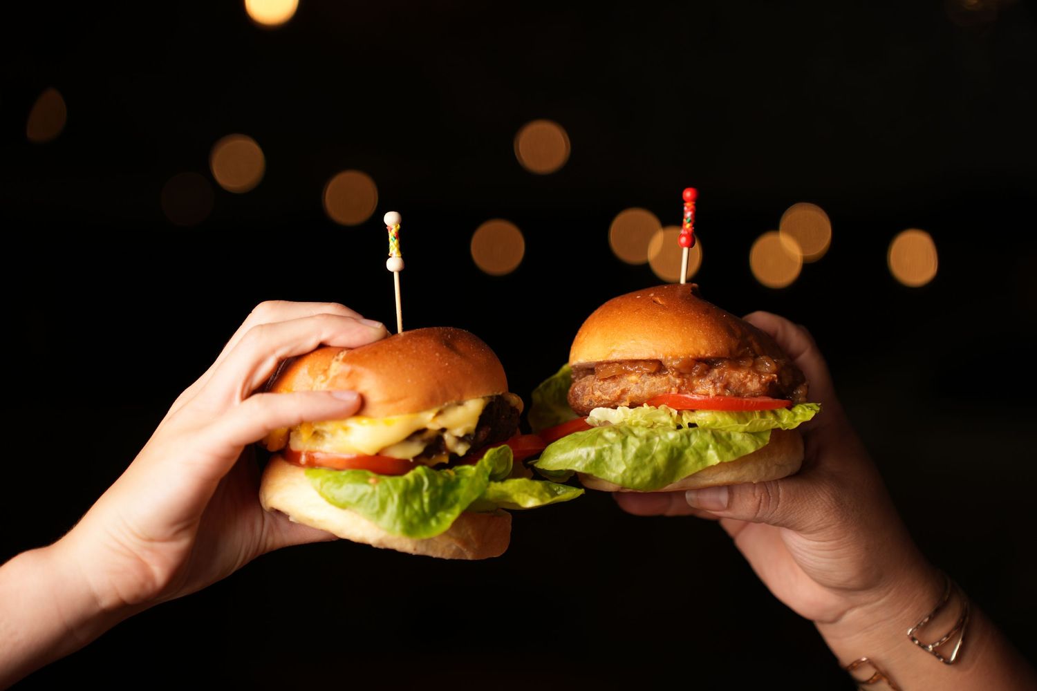Two juicy hamburgers with lettuce held up against dark background with bokeh lights.