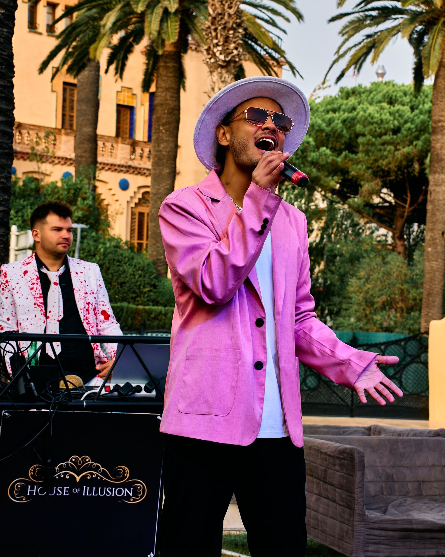 Performer in pink blazer and purple hat singing passionately at an outdoor venue with palm trees and keyboard player.