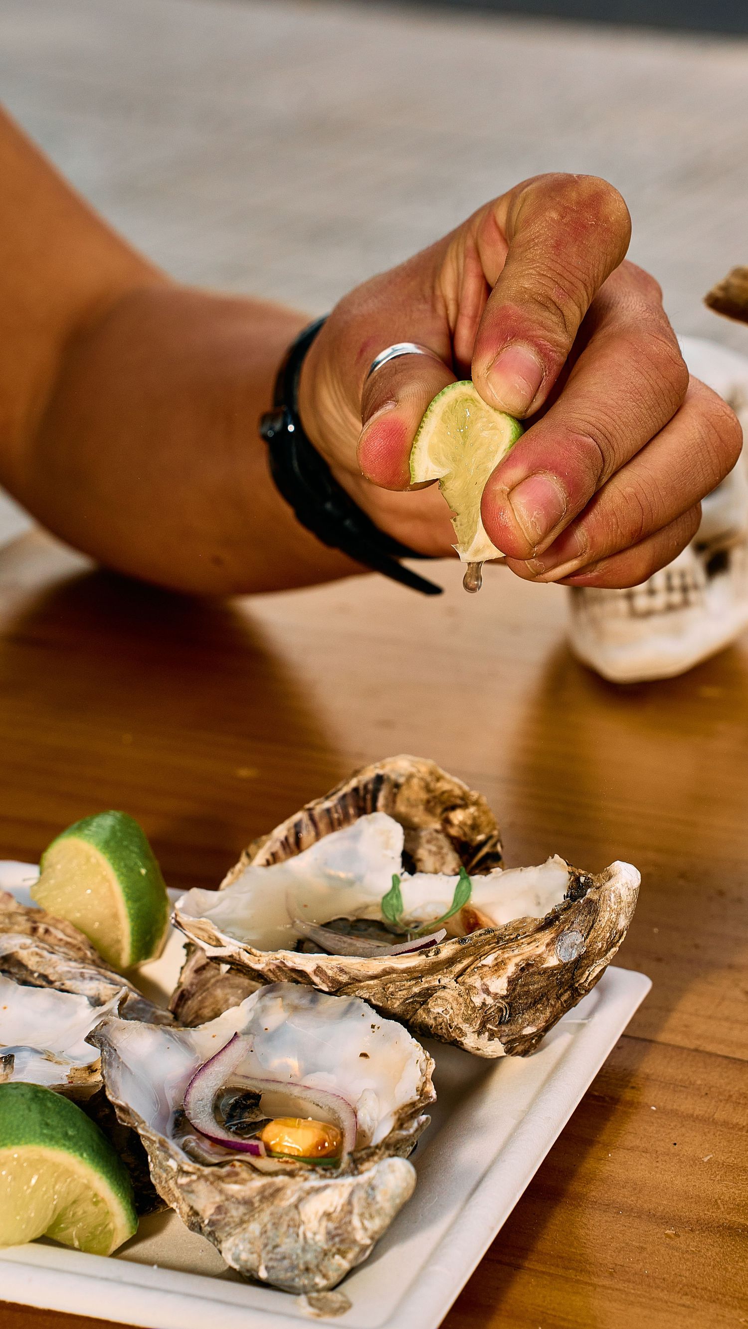 A hand drizzling sauce onto fresh oysters on a white plate with lime wedges on a wooden table.