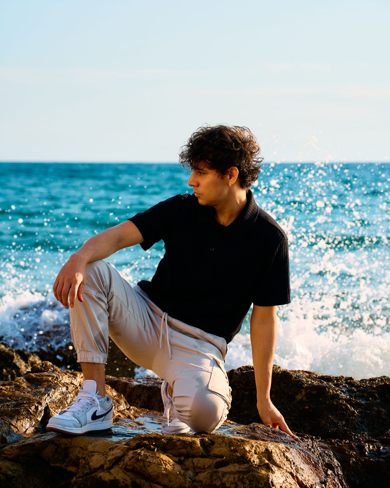 Person relaxes on rocky shore wearing casual attire with bright blue ocean waters in the distance.
