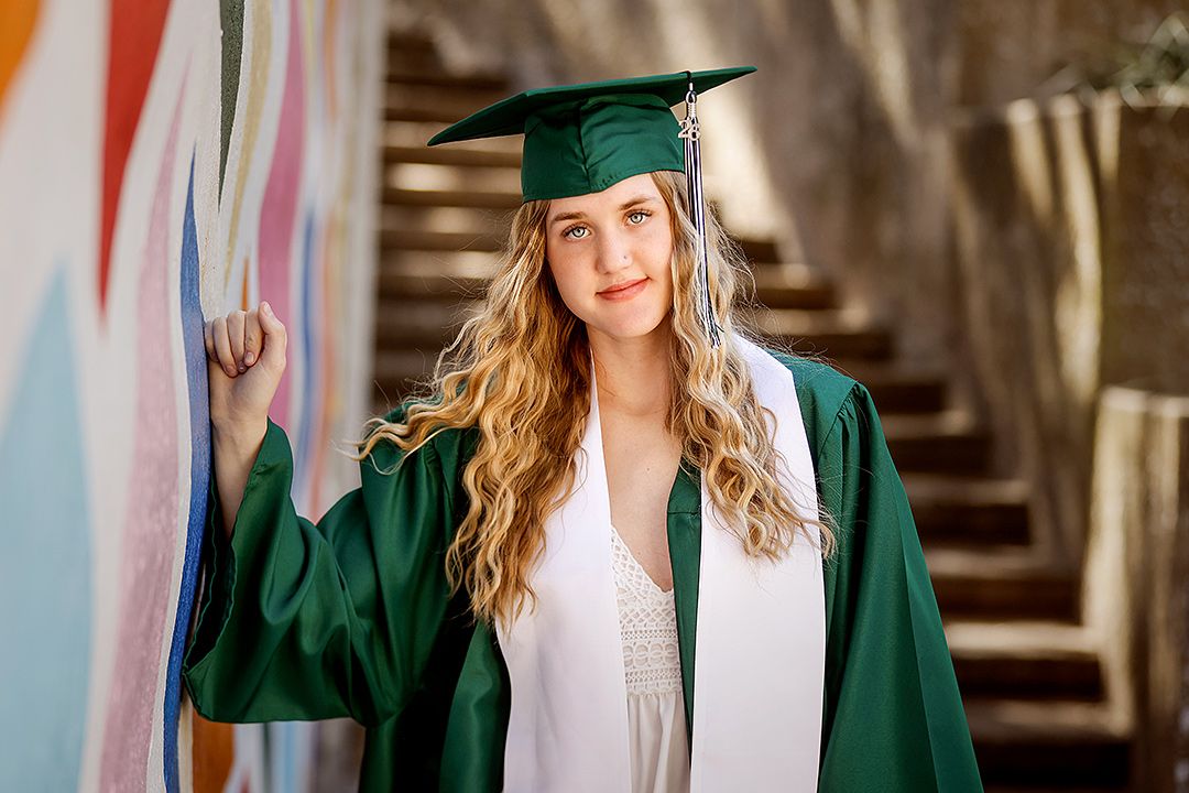 Young woman in green graduation cap and gown smiling beside colorful mural, celebrating academic achievement.