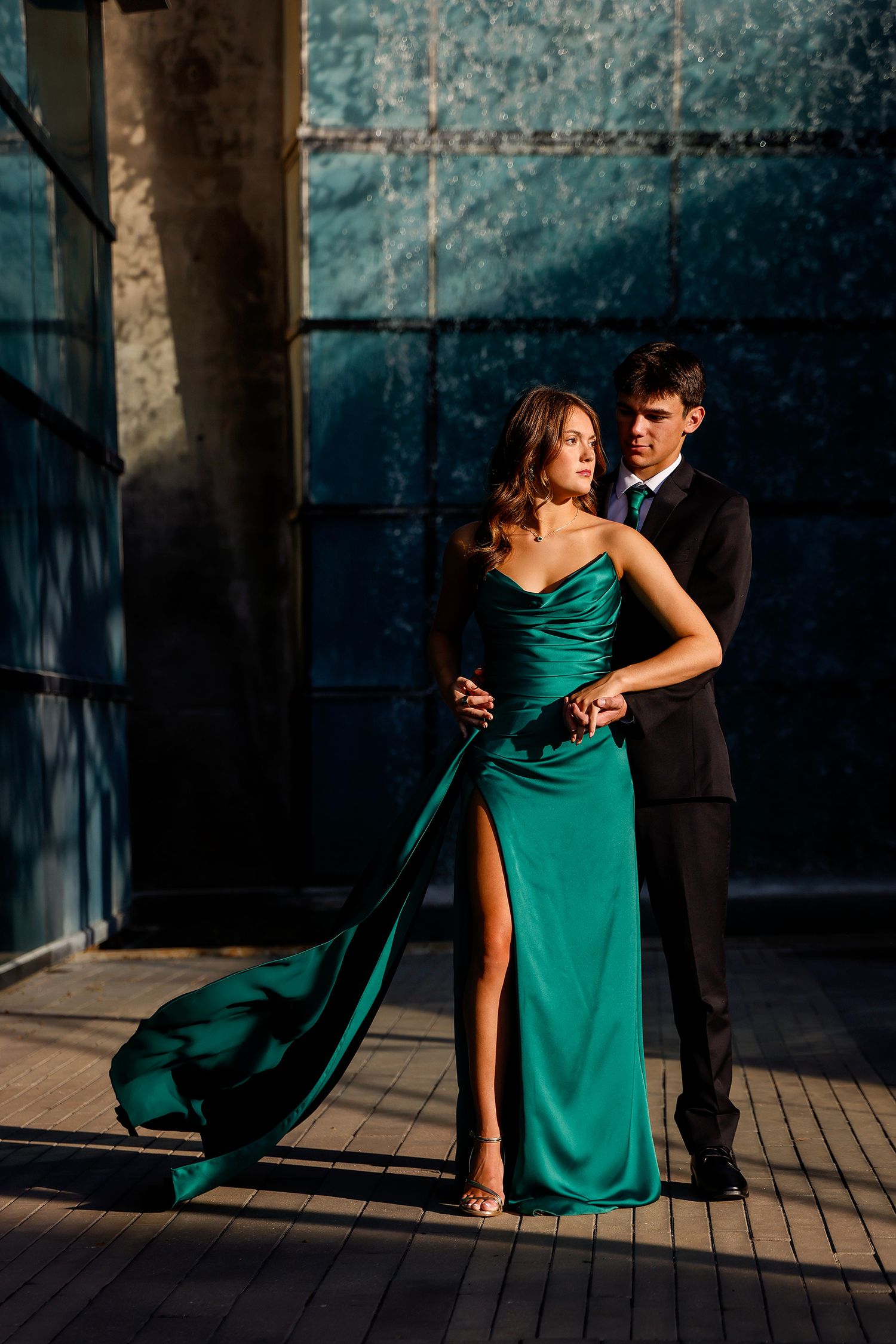 Elegant couple posing outdoors, woman in emerald green satin gown with slit, man in black suit, dramatic lighting.