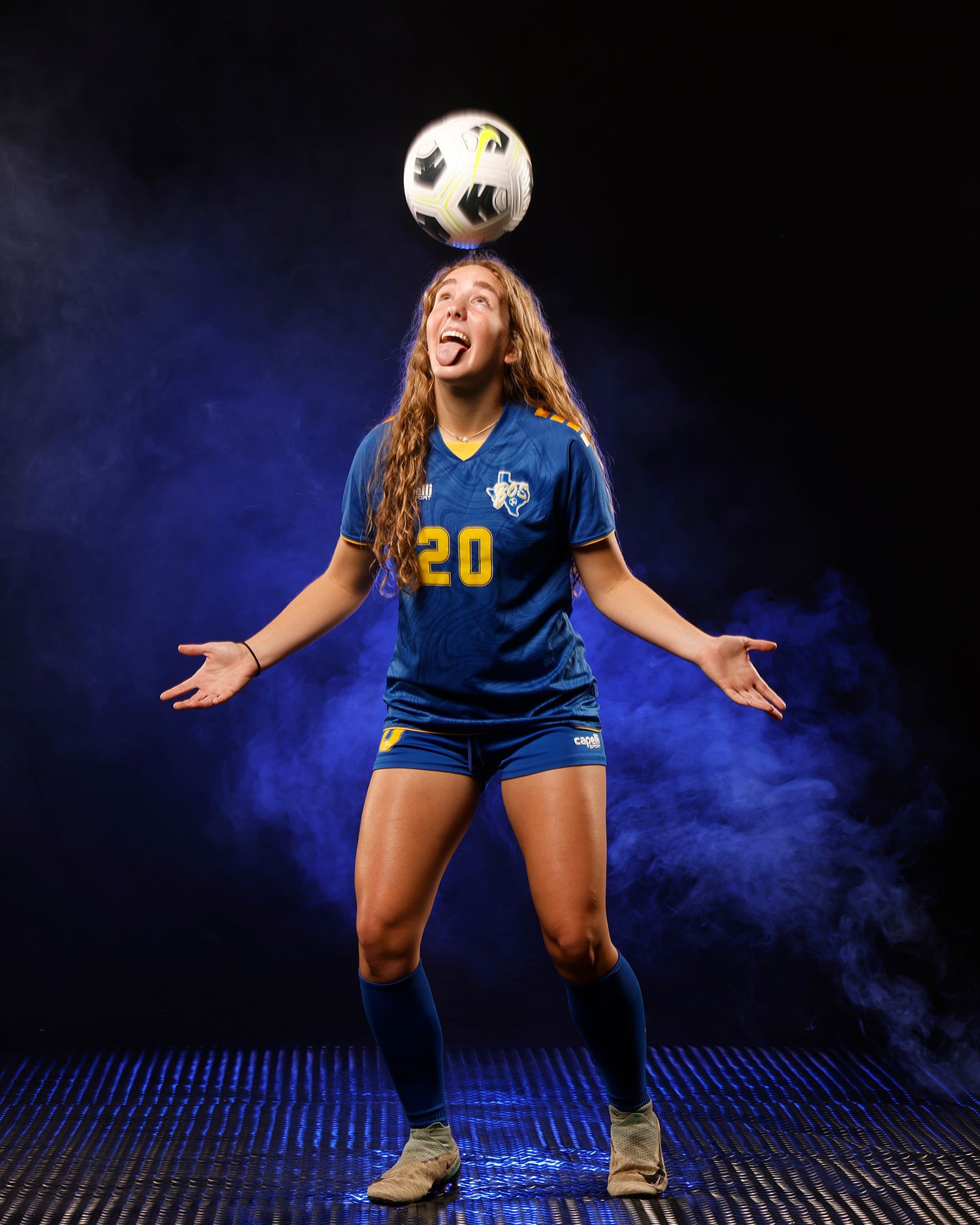 Soccer player #20 balances ball on head with arms out in blue smoky studio.