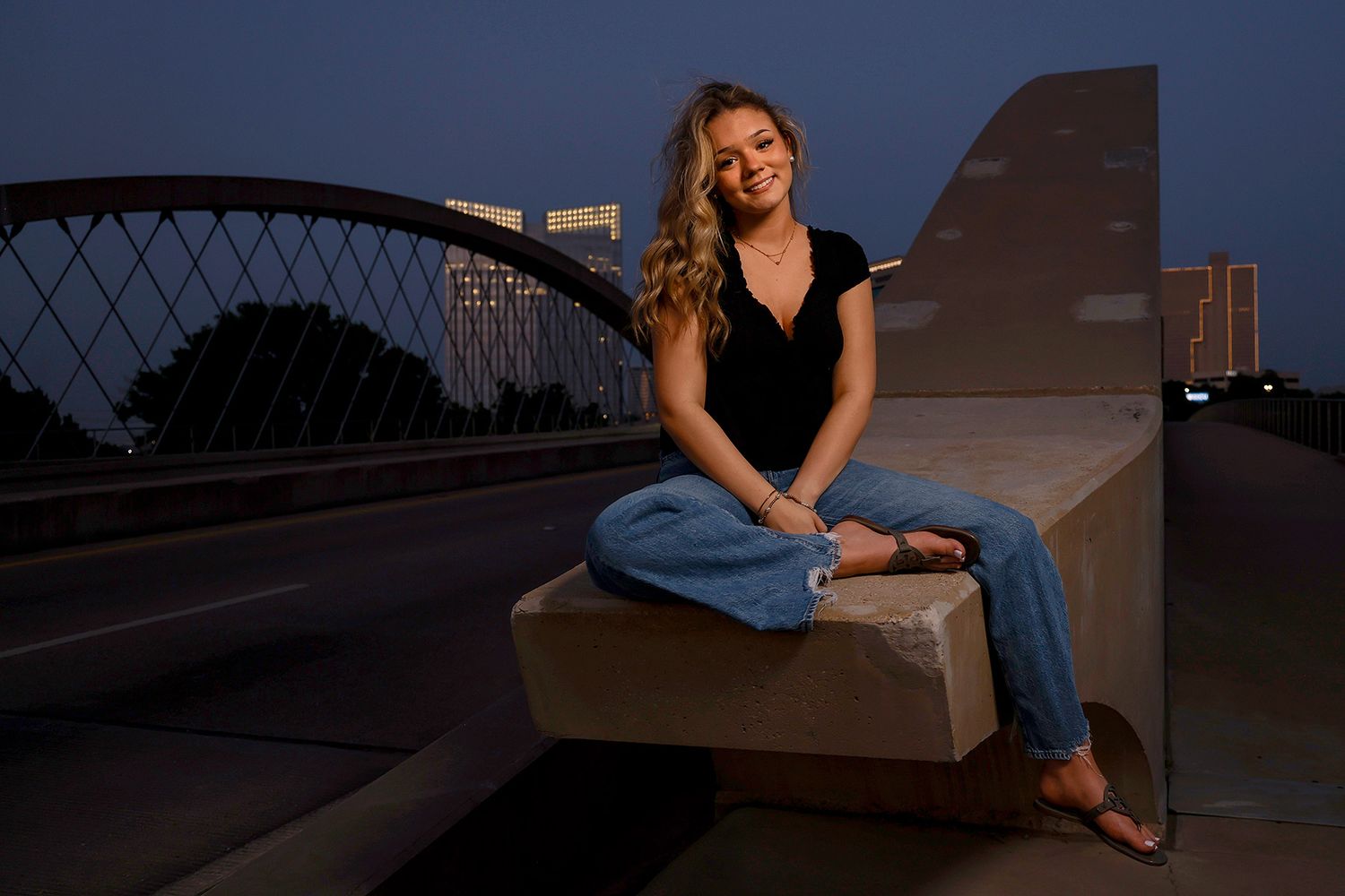 Young woman in black top and jeans sits on a concrete ledge near an arched bridge at dusk.