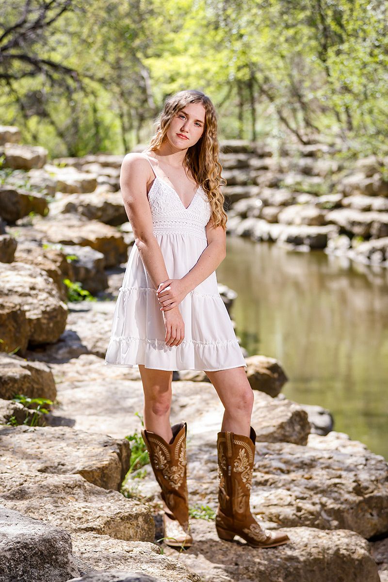 Young woman in white sundress and cowboy boots poses confidently on rocky creek bank outdoors.