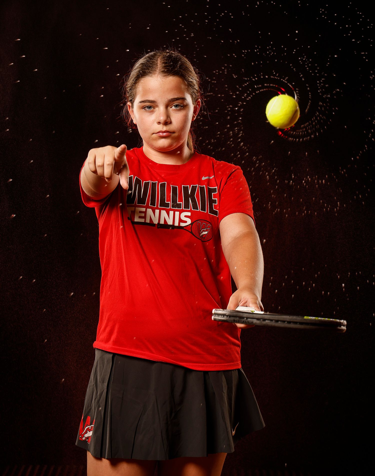 Young female tennis player in red Willkie Tennis shirt poses with racket and ball in action shot.