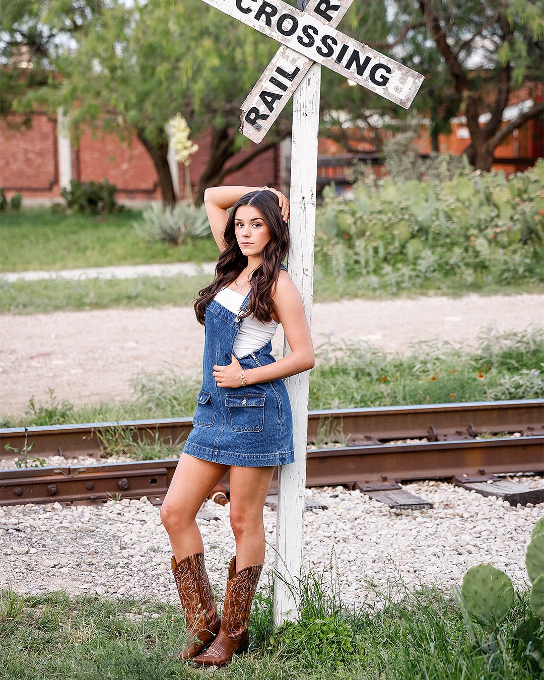 Young woman in denim overalls dress and cowboy boots poses by a railroad crossing sign on train tracks.