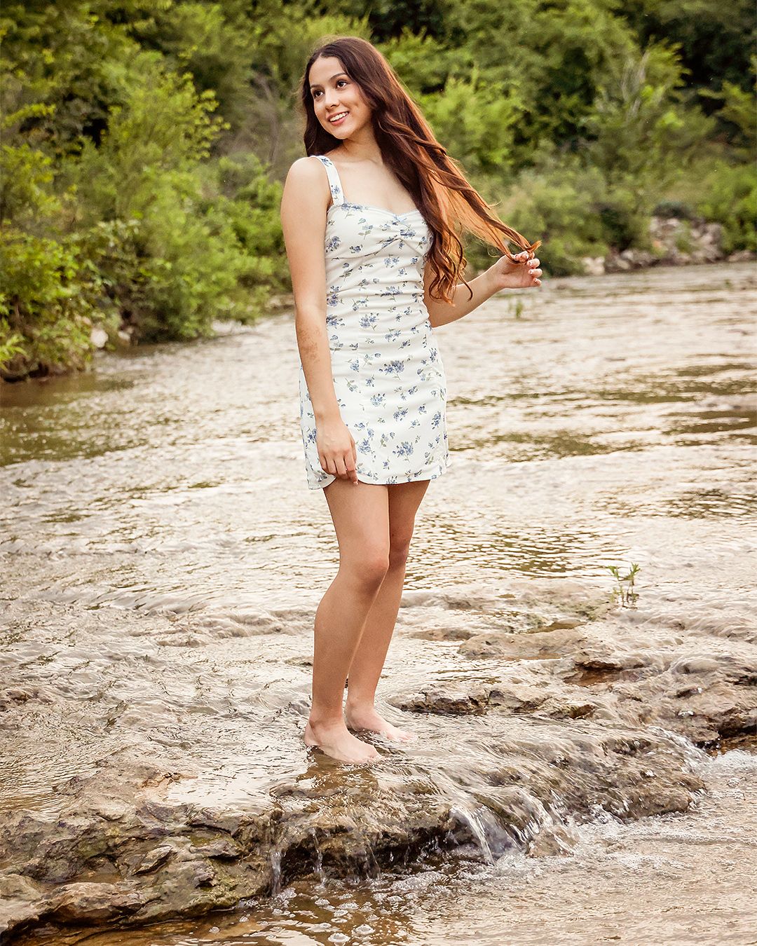 Young woman in floral mini dress standing barefoot on rocks in a flowing creek surrounded by lush greenery.