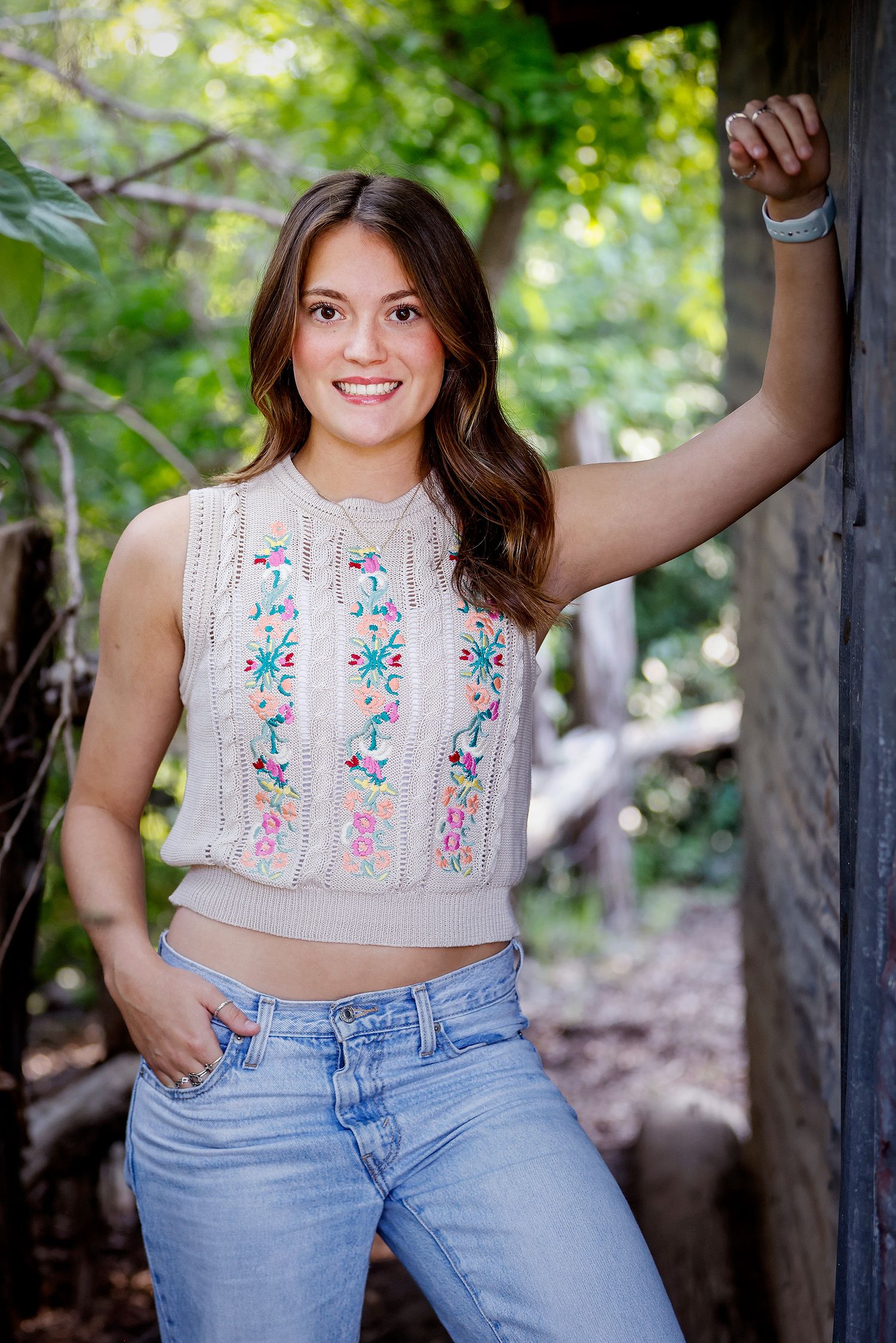 Young woman smiling in embroidered crop top and jeans, leaning against rustic wooden post outdoors.