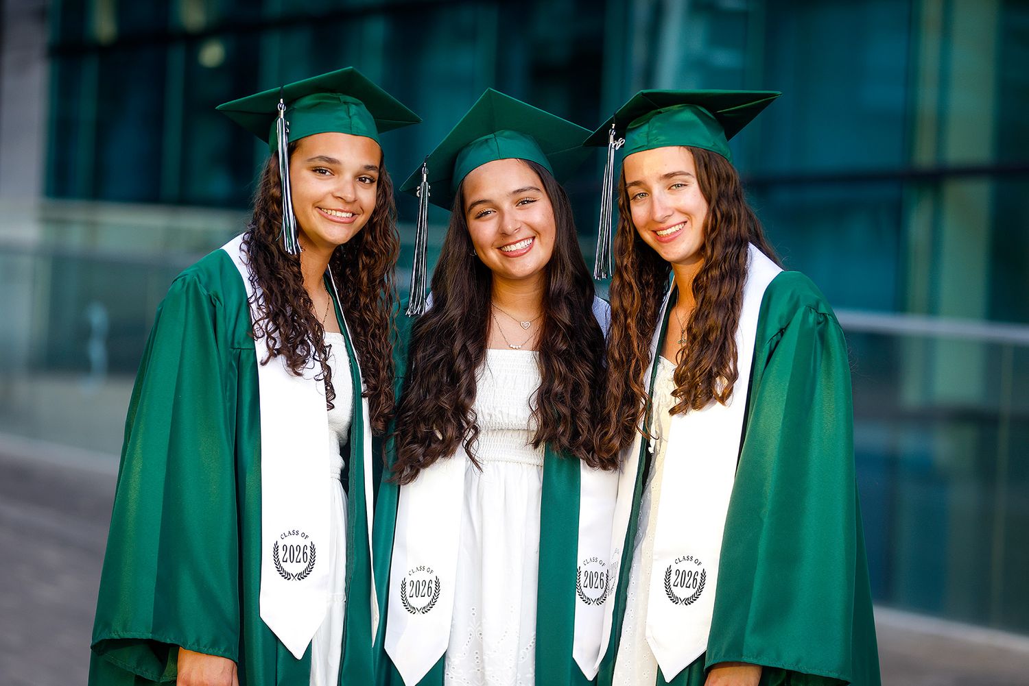 Three smiling graduates in green caps and gowns posing together at an outdoor graduation ceremony.