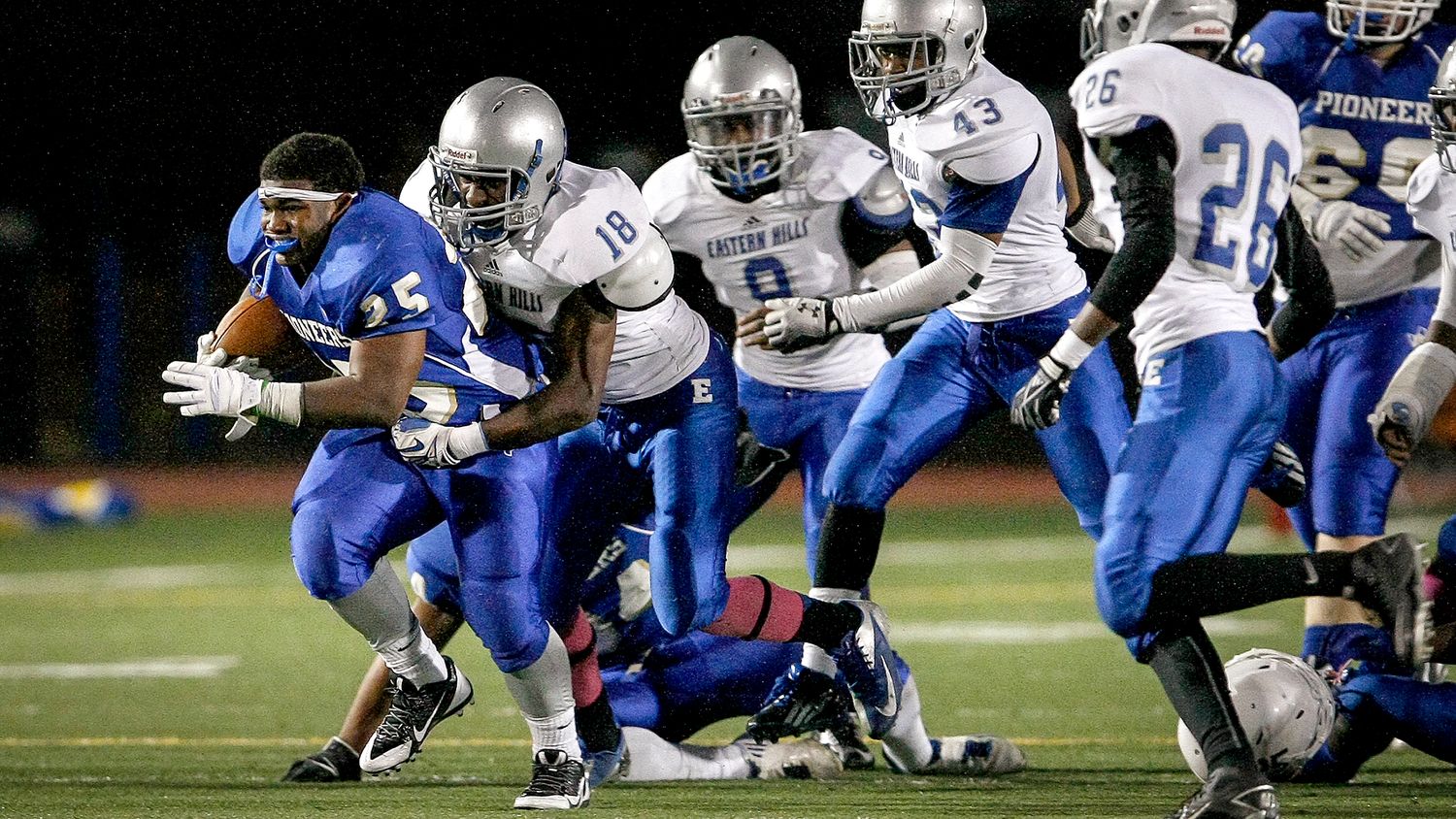 High school football running back breaks through defensive line during competitive night game.