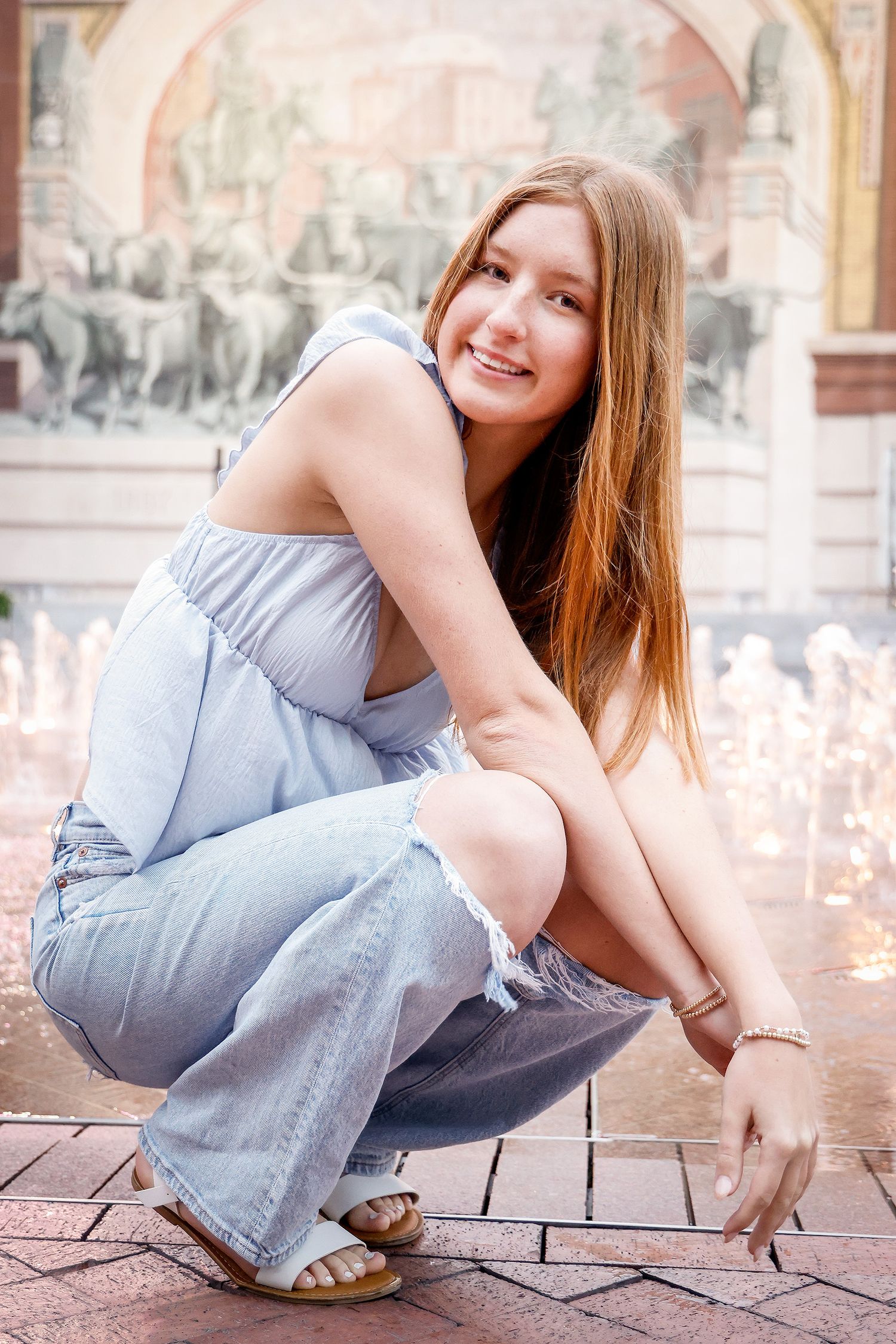 Young woman in blue outfit crouching near a fountain, smiling outdoors near a decorative mural backdrop.