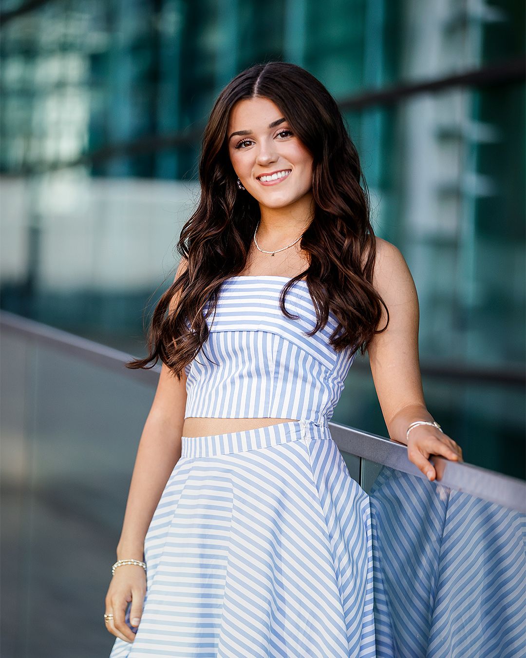 Young woman in blue striped crop top and skirt smiling outdoors near glass building railing.