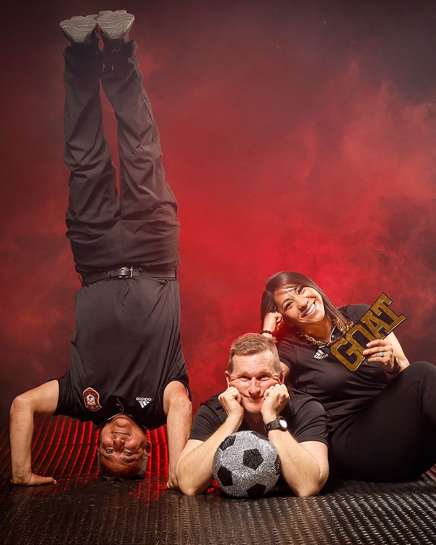 Three sports coaches pose playfully with a soccer ball, one doing a handstand, against a red smoky backdrop.