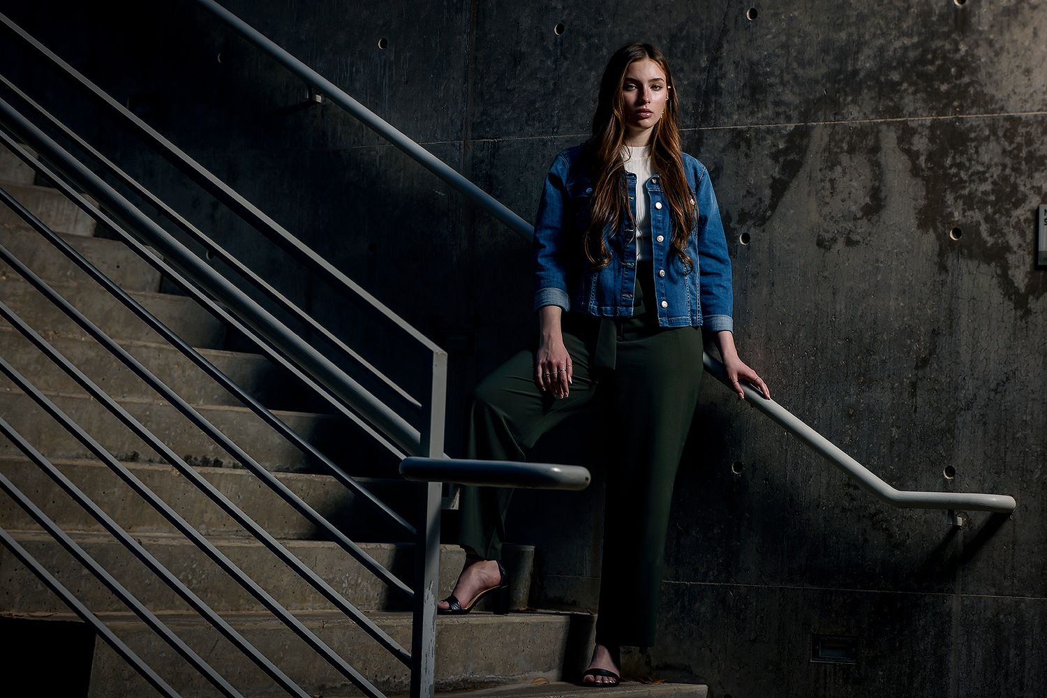 Young woman in blue denim jacket and green pants posing on concrete stairs with metal railing.