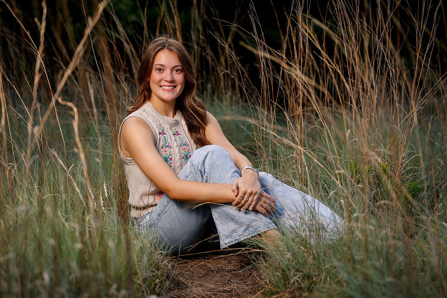 Young woman with long hair sits smiling in tall golden grass, wearing floral top and jeans, outdoor portrait.