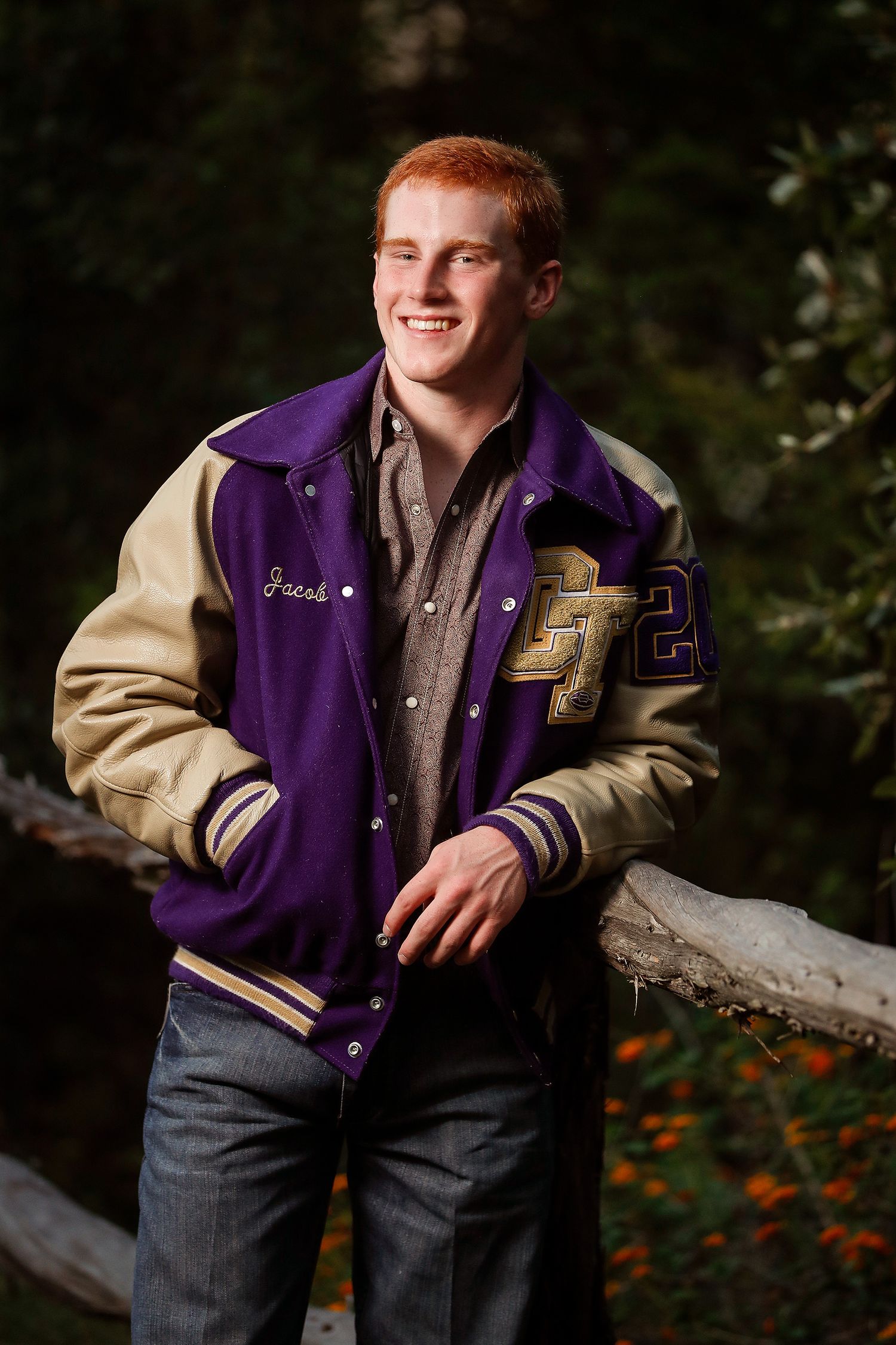 Smiling teenage boy with red hair wearing a purple and gold varsity jacket with name Jacob, posing outdoors.