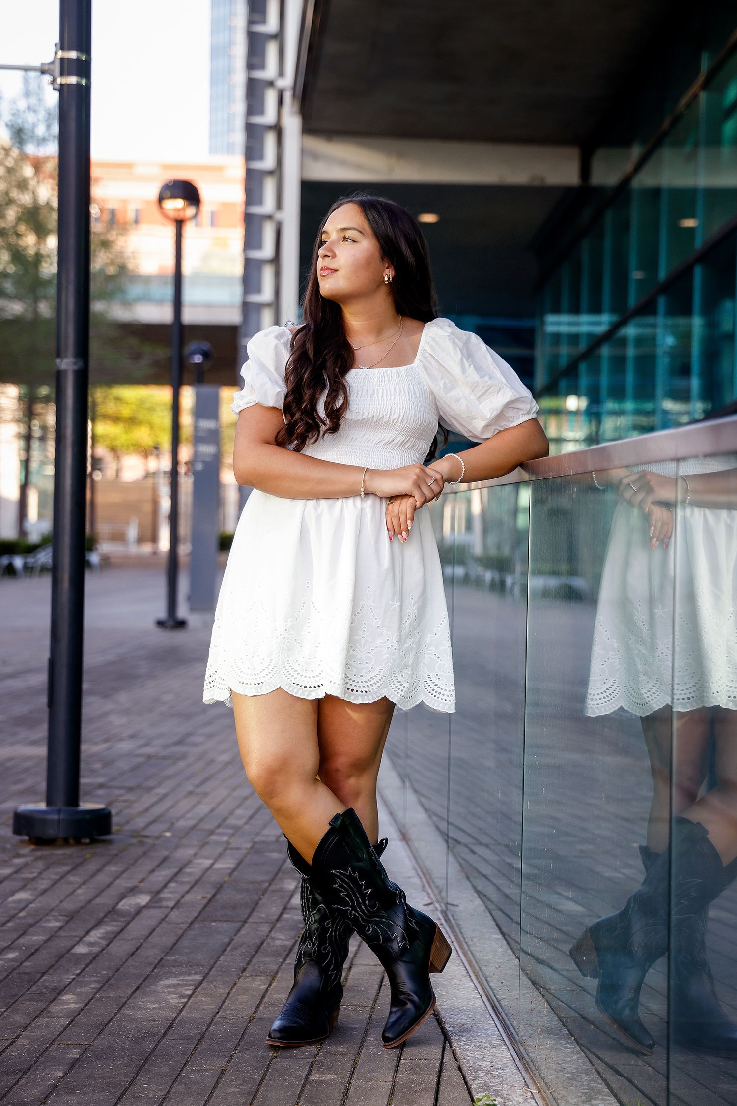 Young woman in white eyelet dress and cowboy boots leaning against glass railing in urban setting.
