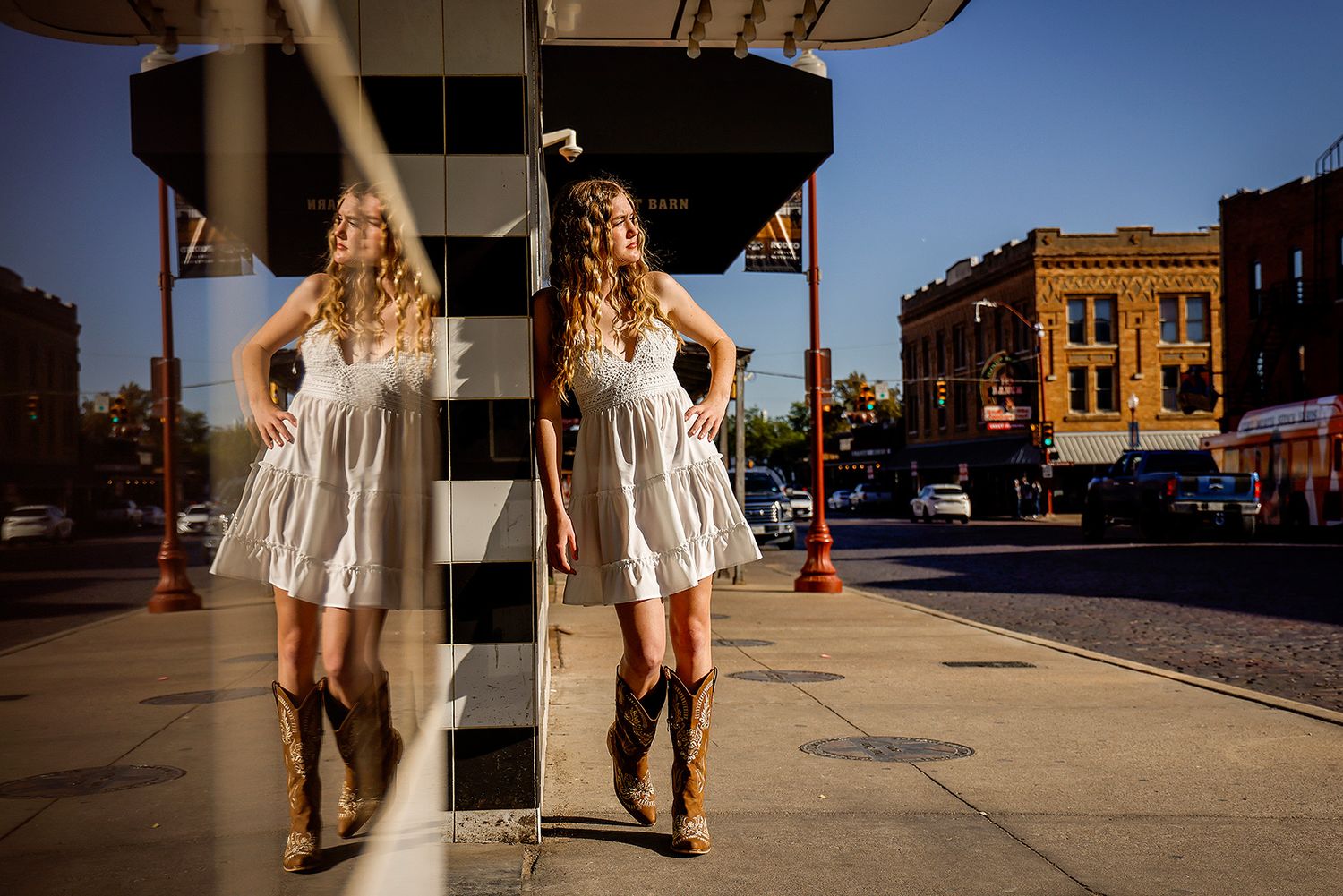 Young woman in white dress and cowboy boots poses with her reflection in a mirrored column on a sunny street.