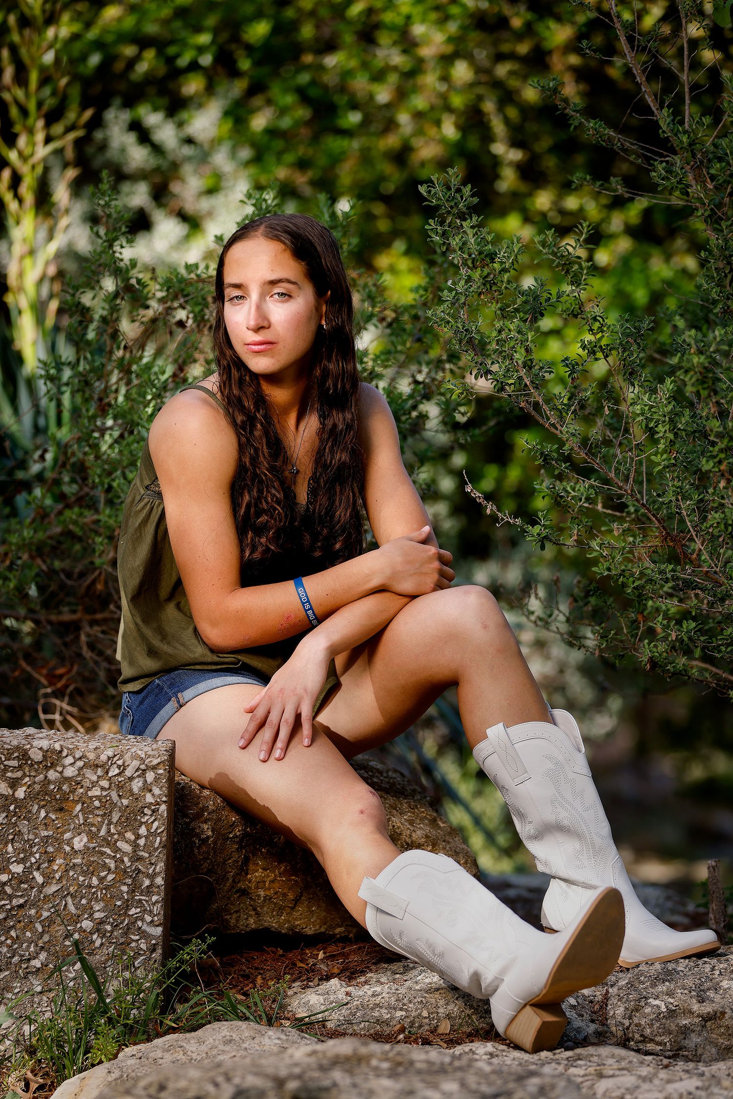 Young woman in denim shorts and white cowboy boots sits on rocks outdoors surrounded by green shrubs.