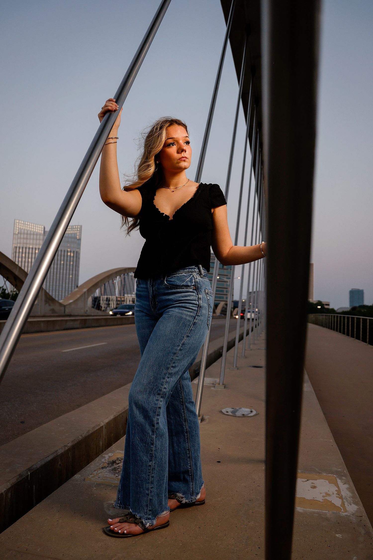 Young blonde woman posing on a bridge at dusk, wearing a black top and wide-leg jeans with city skyline behind her.