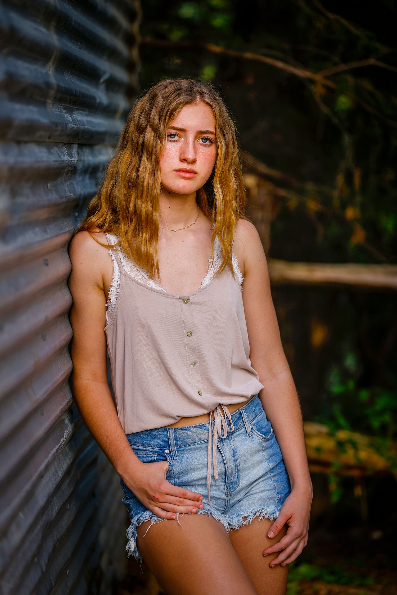 Blonde teenage girl in beige tank top and denim shorts leans against corrugated metal wall.