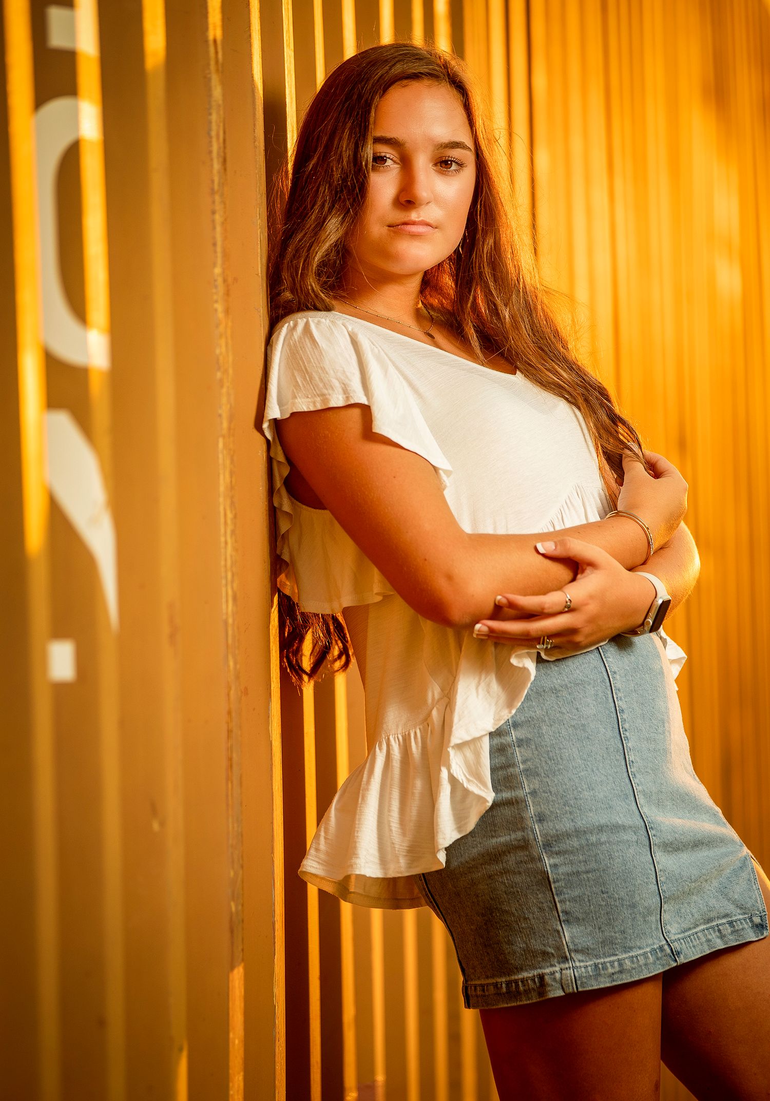 Young woman in white top and denim skirt poses against golden yellow corrugated wall outdoors.