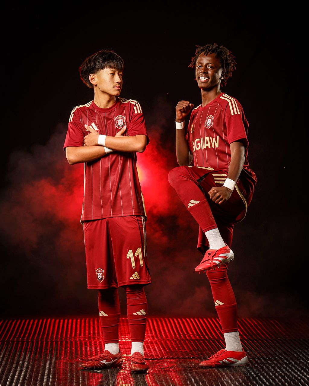 Two male soccer players in red uniforms strike dynamic poses against a dramatic dark smoky background.