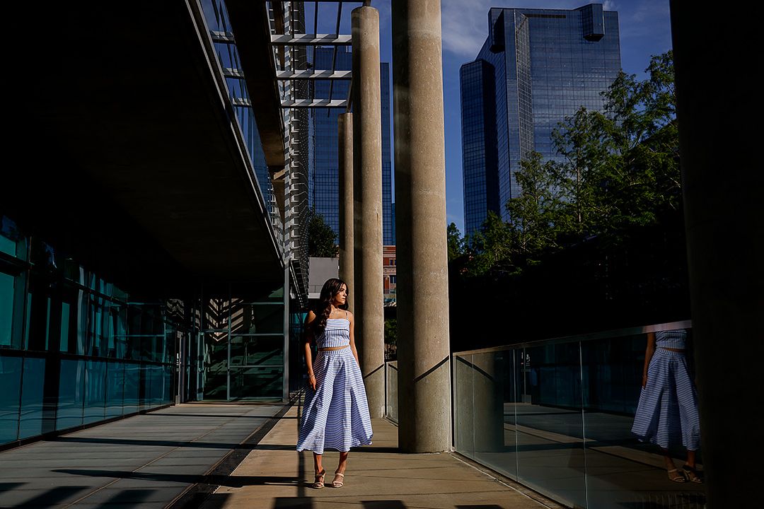 A woman in a white dress stands between modern glass buildings and stone columns in an urban setting.
