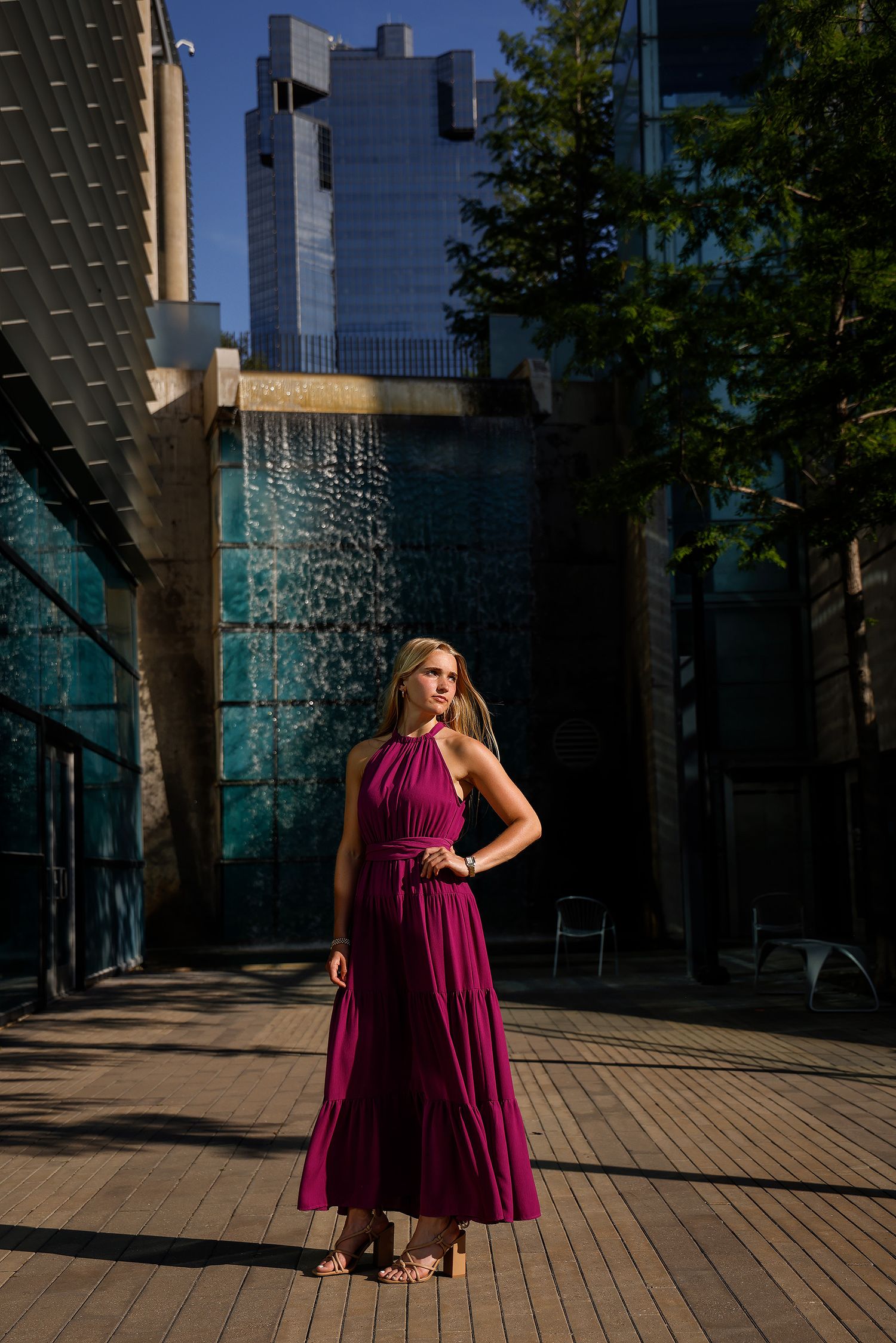 Blonde woman in magenta maxi dress poses near urban waterfall fountain with glass skyscrapers and trees in background.
