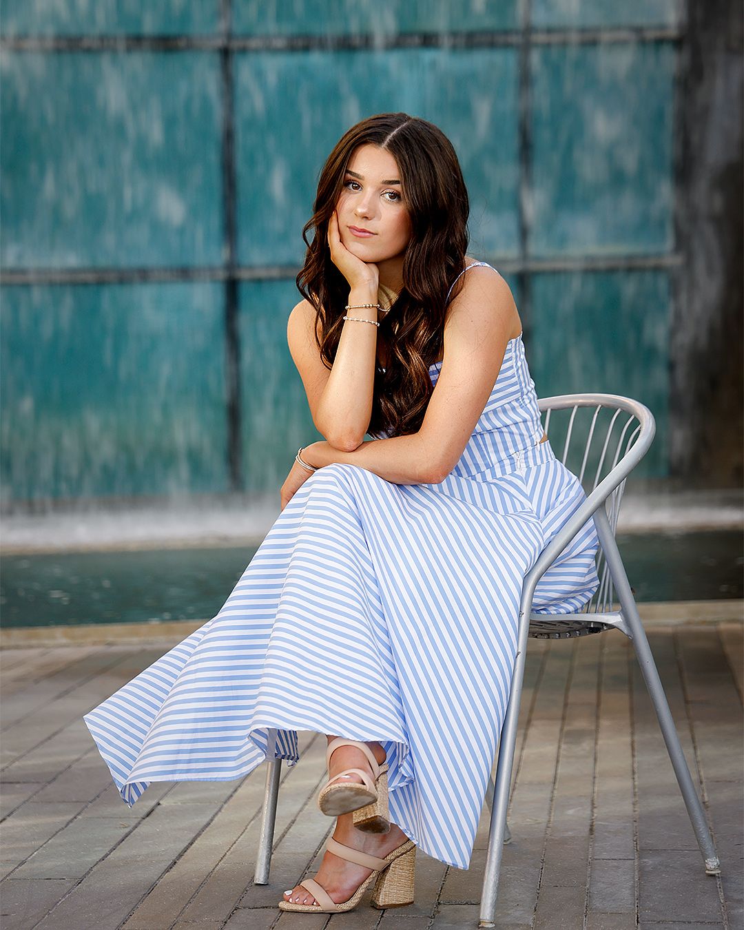 Young woman in blue striped maxi dress seated on white chair near teal fountain wall.