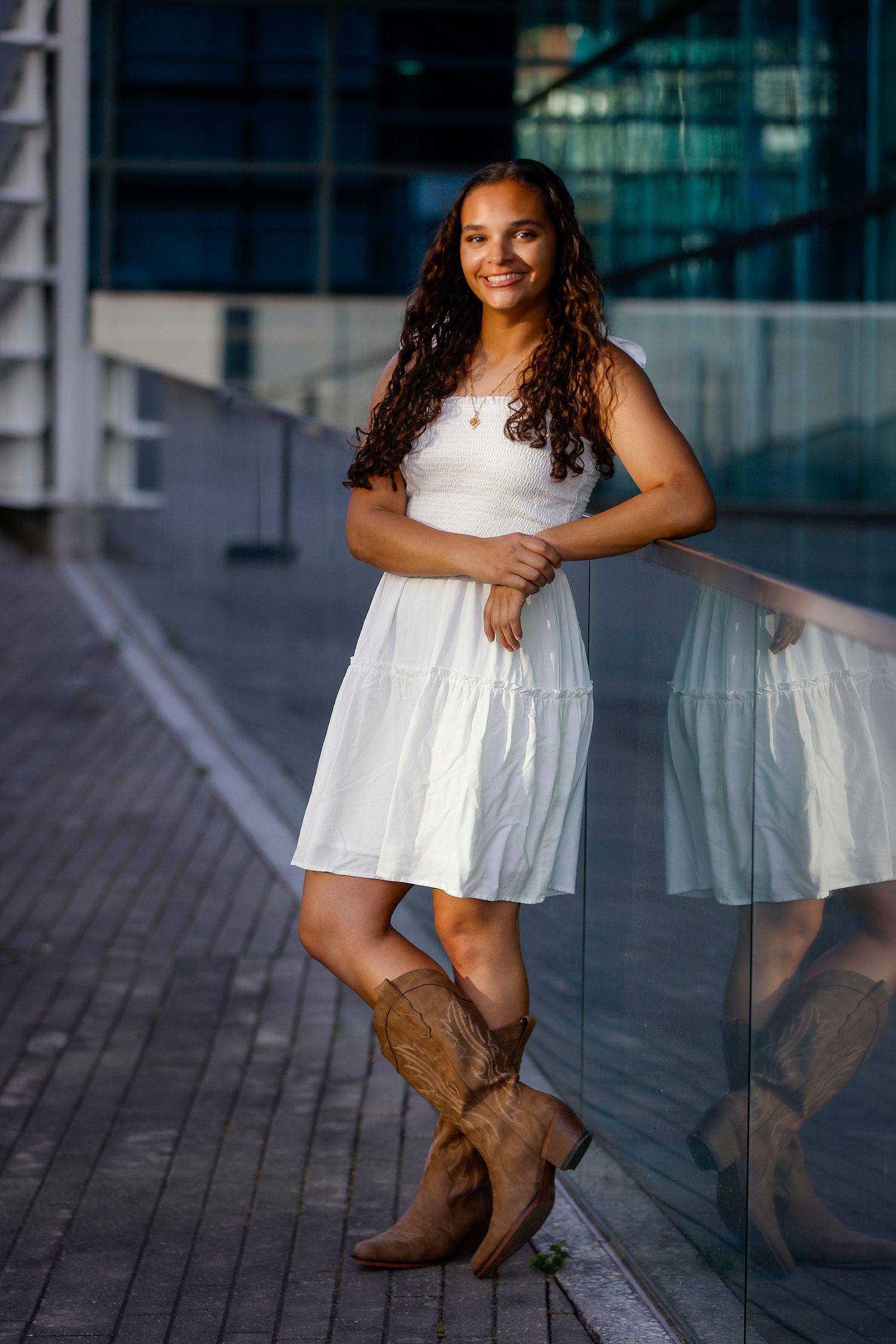 Young woman in white dress and cowboy boots leaning on glass railing, smiling outdoors in urban setting.