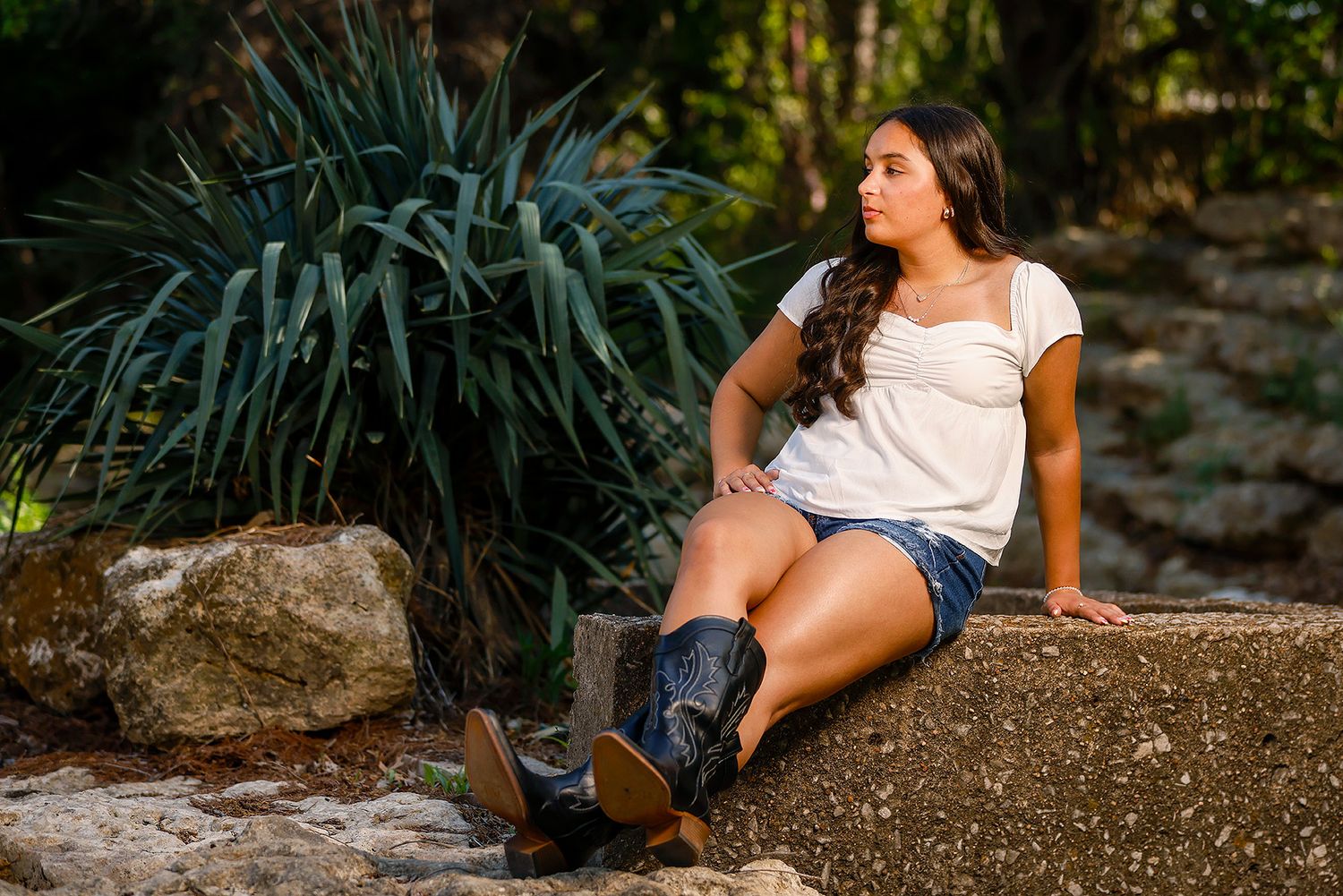Young woman in white top, denim shorts, and cowboy boots sitting on rocks surrounded by lush greenery.