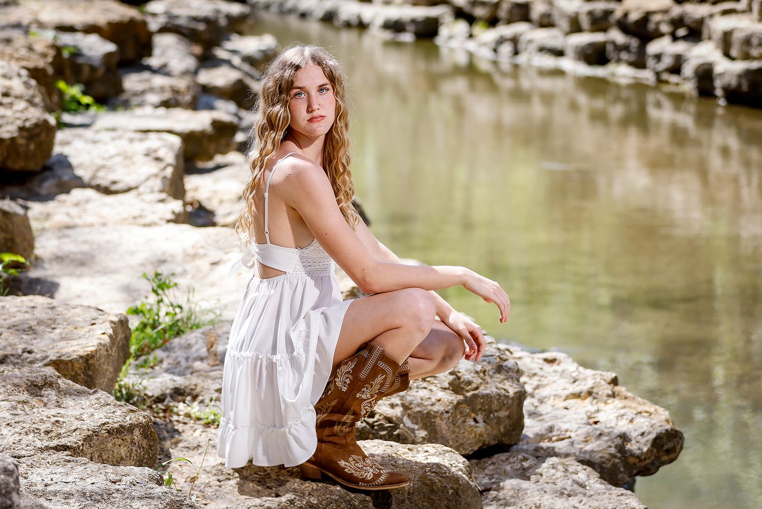 Young woman in white dress and cowboy boots sits gracefully on rocks beside a serene creek.