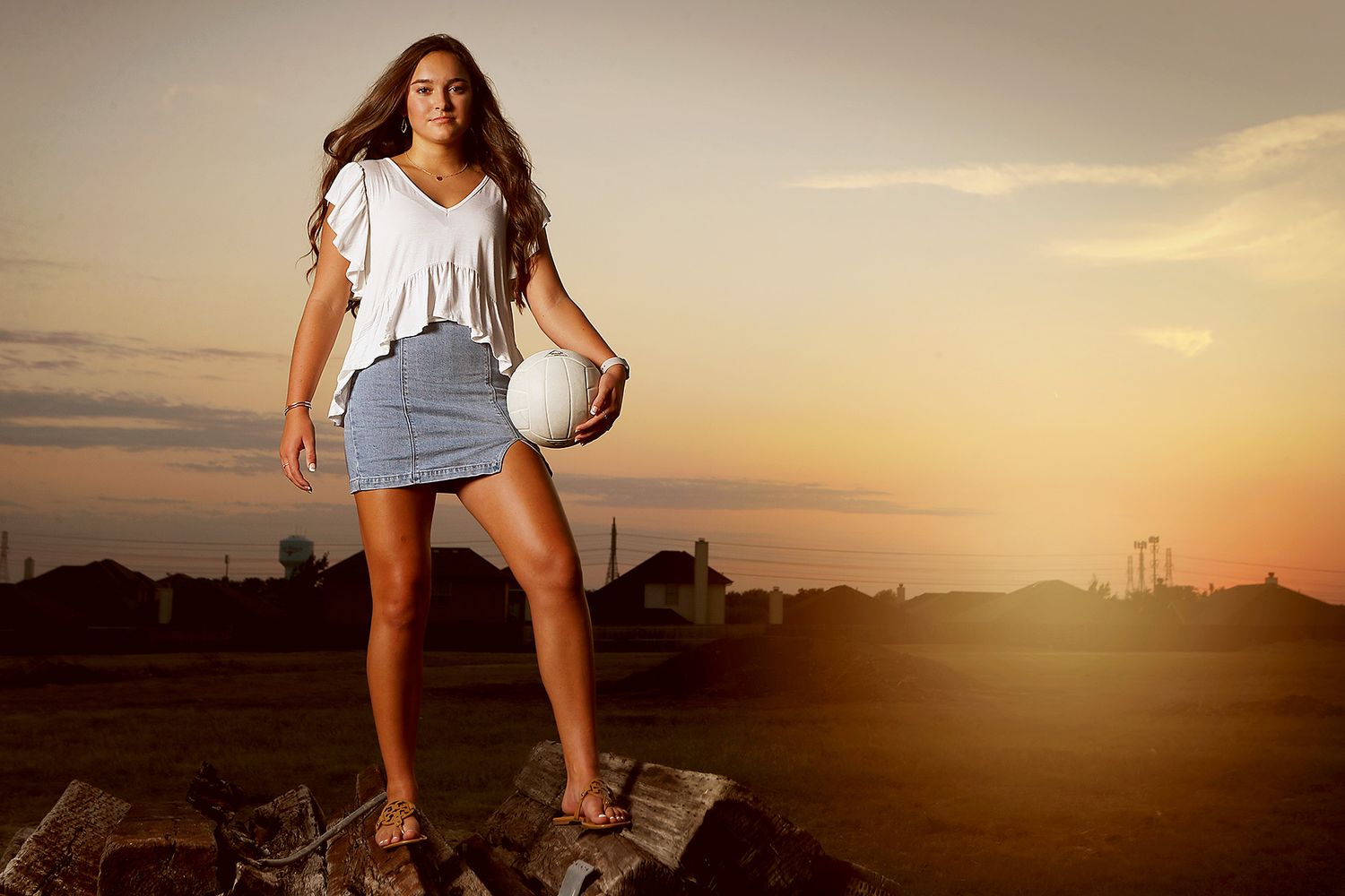Young woman in white top and denim skirt holding a volleyball at sunset in an outdoor setting.