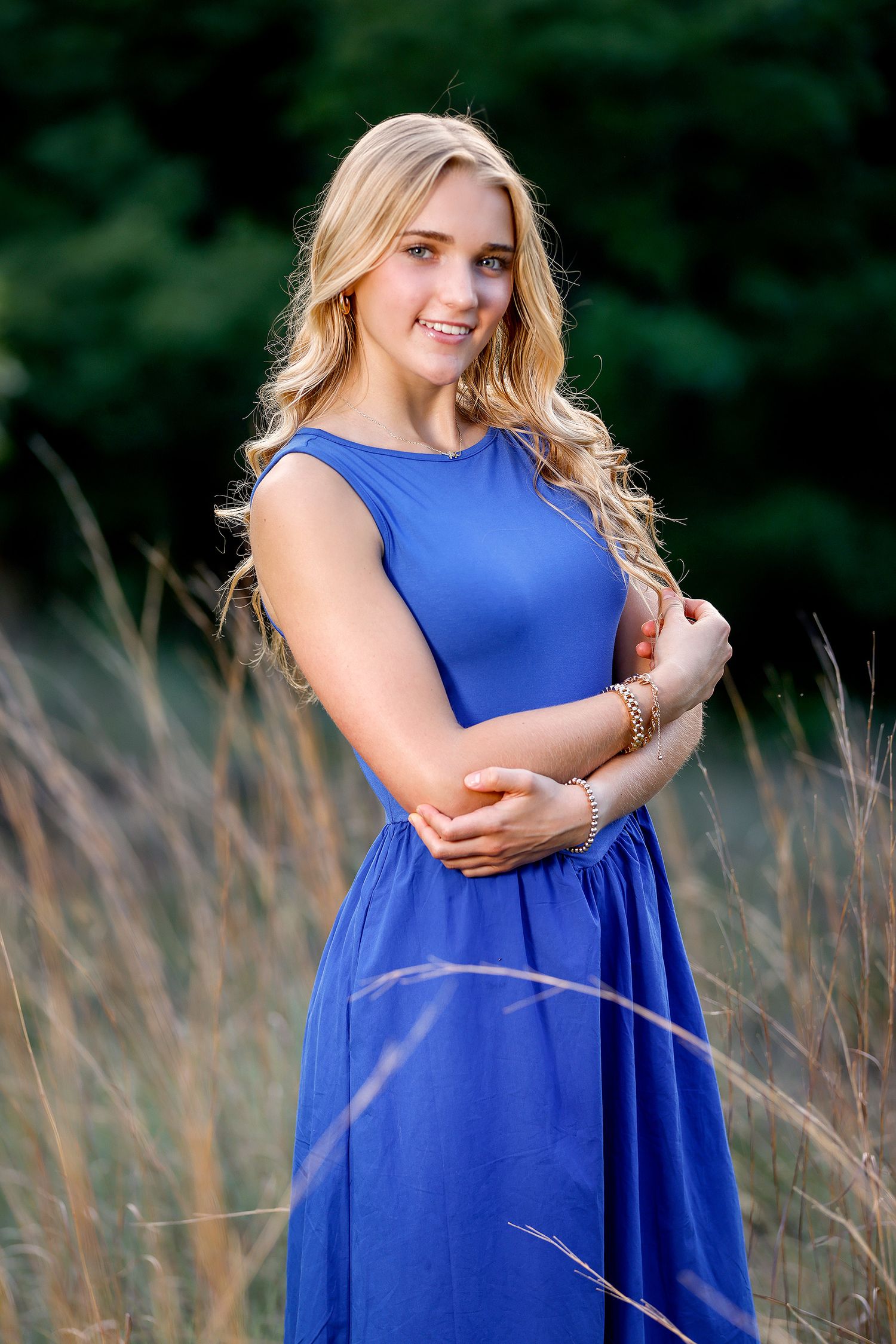 Young blonde woman in a royal blue maxi dress smiling outdoors in a field of tall grass with green trees behind her.