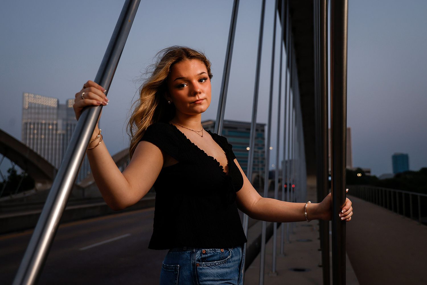 Young woman in black top and jeans poses on a bridge at dusk, holding metal railings with city skyline behind her.
