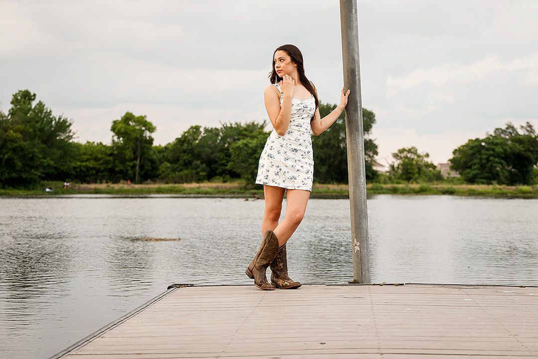 Young woman in white floral mini dress and cowboy boots posing on a wooden dock by a lake.
