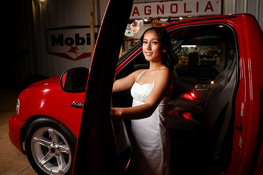 A young woman in a white dress poses elegantly inside a red car near vintage Mobil and Magnolia gas station signs.