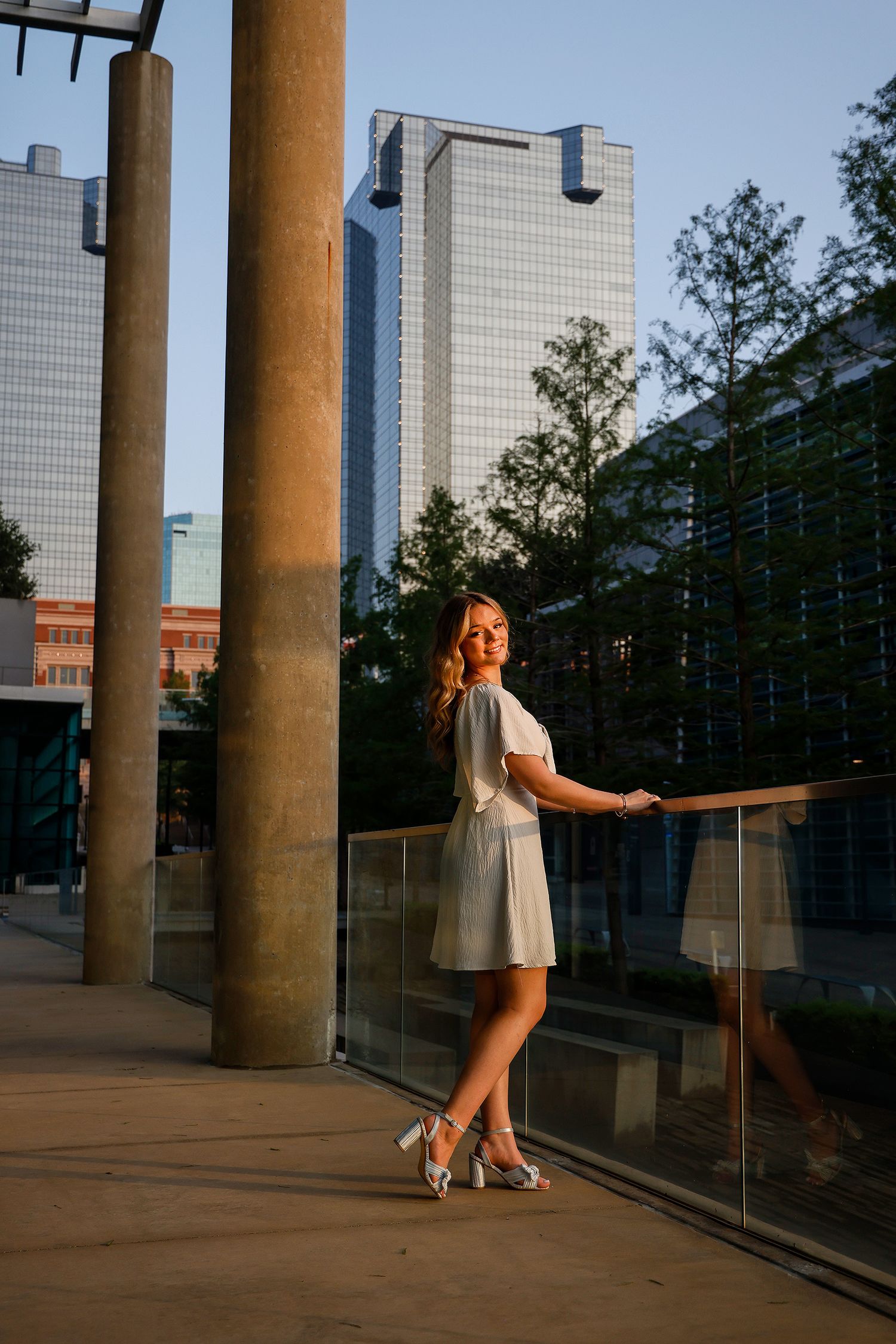 Young woman in white dress and heels poses by glass railing with city skyscrapers in golden hour light.