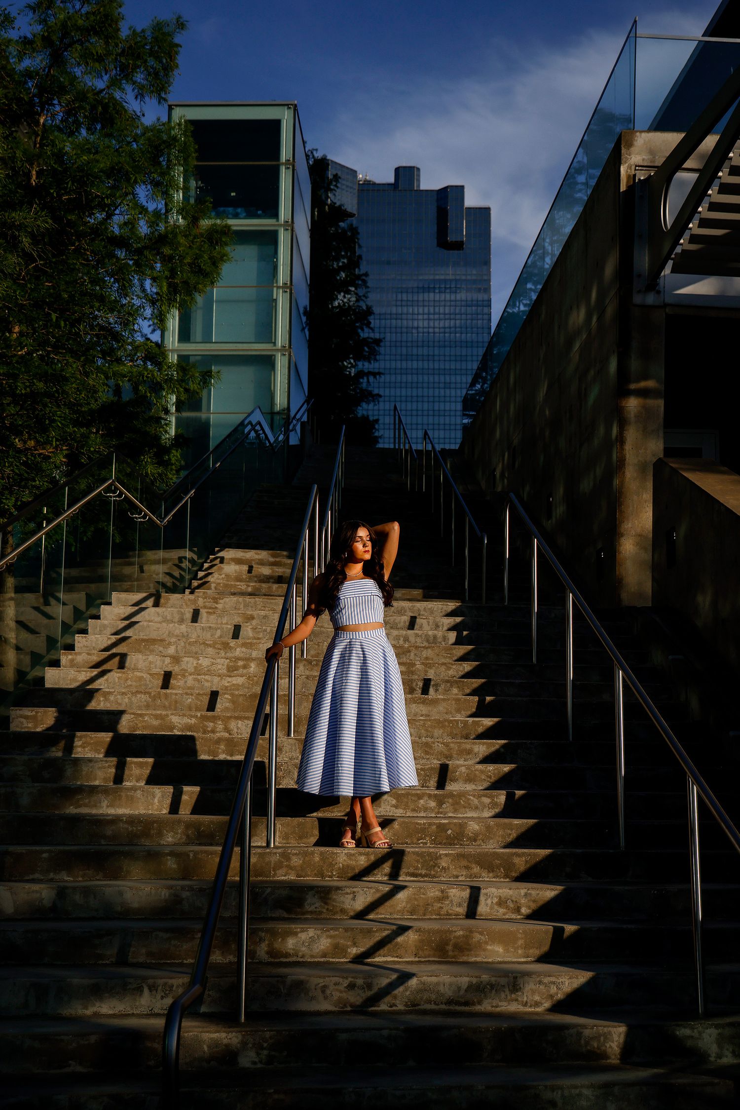 Young woman in blue crop top and midi skirt poses gracefully on urban stone steps with modern glass buildings behind her.