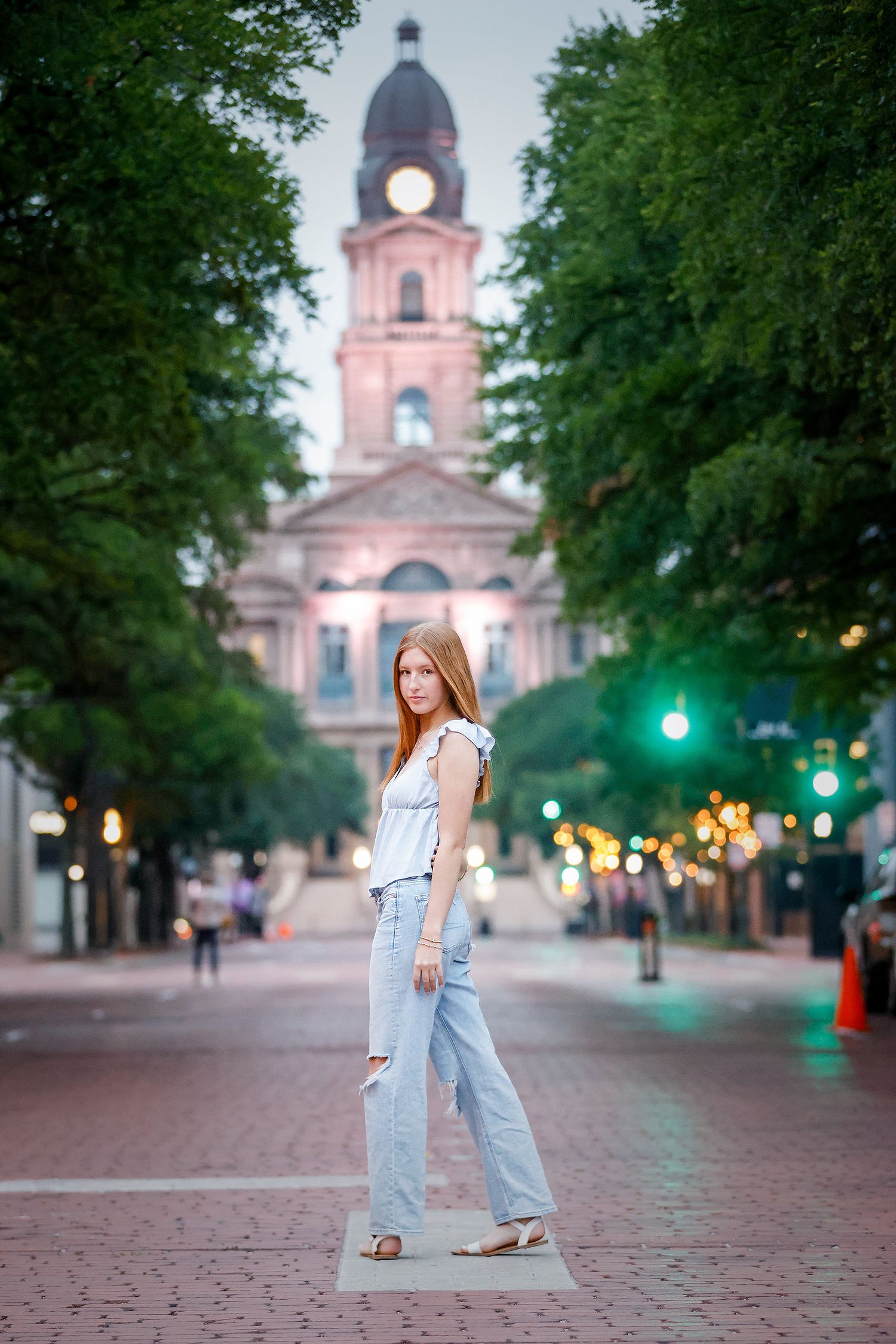 Young redhead woman in ripped jeans and white top poses on brick plaza before historic clock tower at dusk.