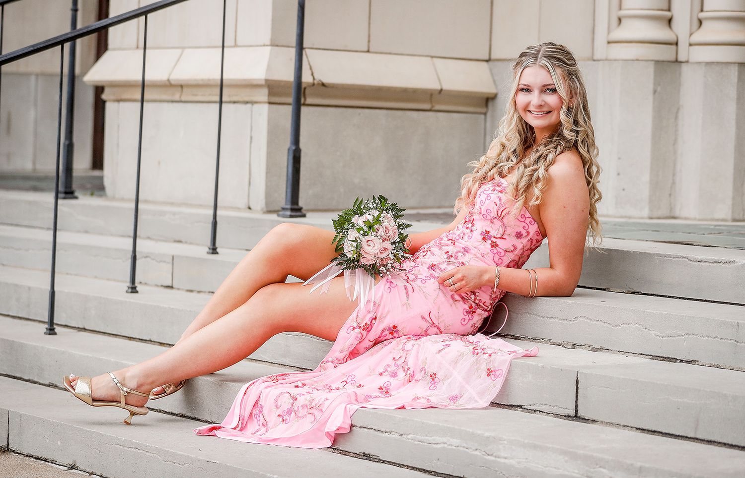 Young woman in pink floral dress sitting on stone steps holding a bouquet of flowers.