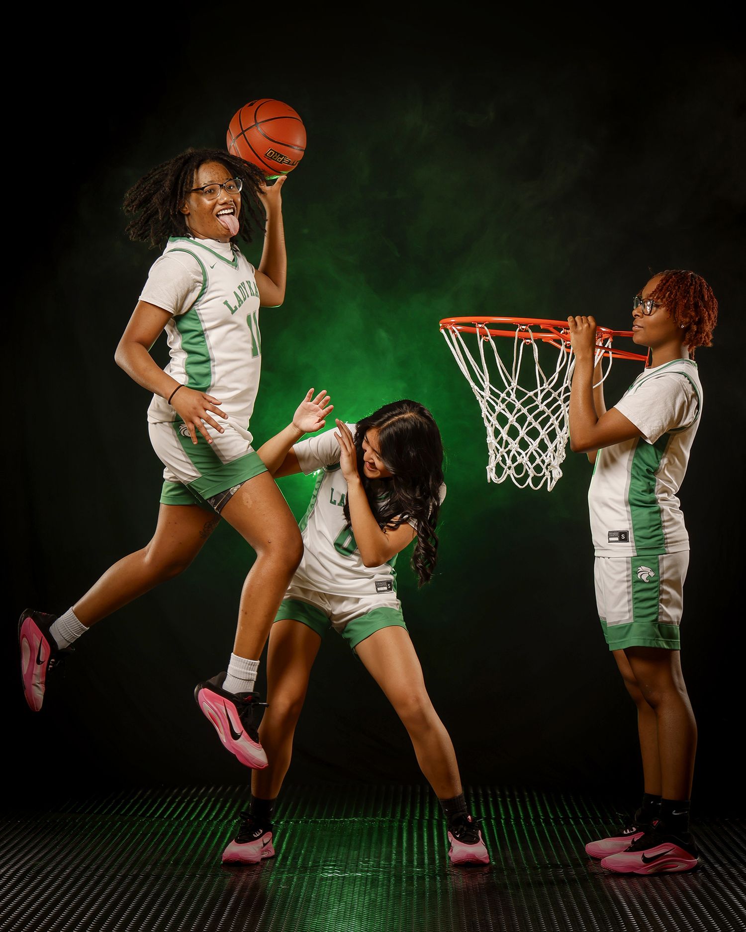 Three female basketball players pose dynamically with hoop in green-lit studio.